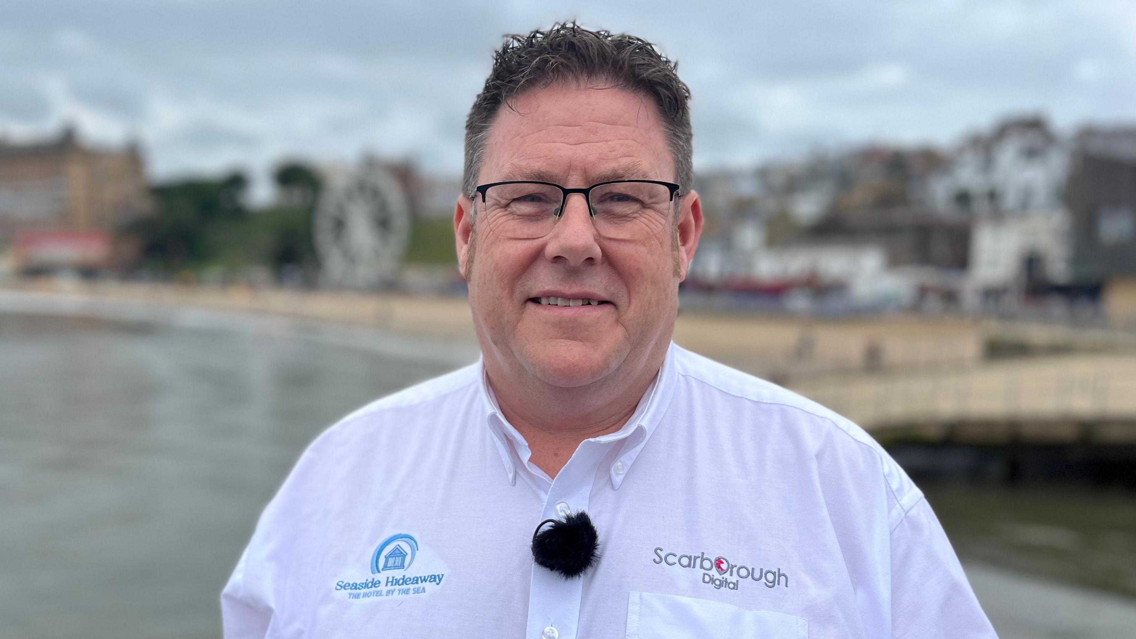 Jim Grieve wears a white shirt and stands by the sea looking at the camera