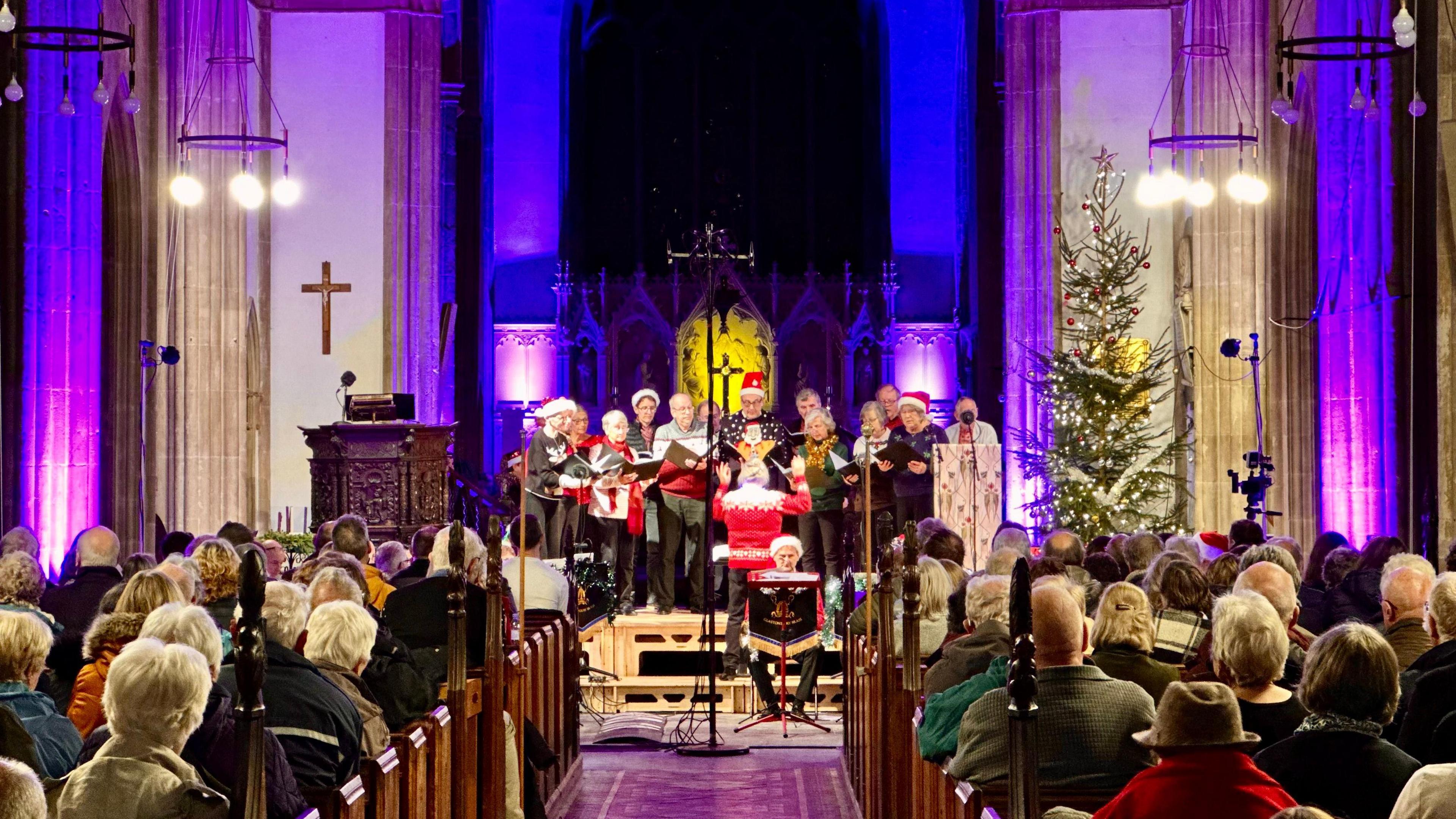 A church with lots of people sat on the seats. There is a choir performing on the stage.