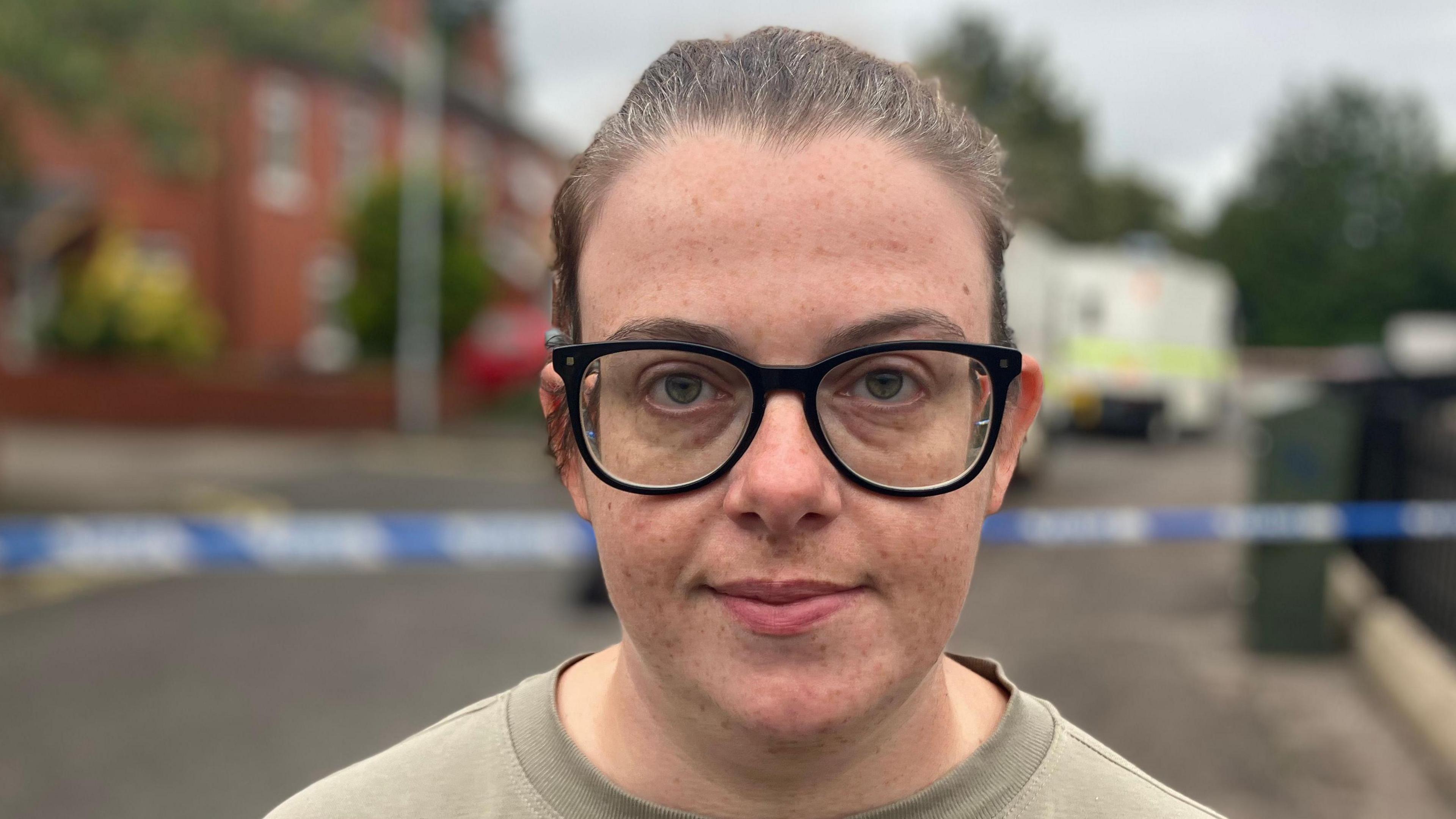 Cllr Ruth Brooks standing by a police cordoned street. Her dark hair is tied up and she is wearing a light jumper and black framed glasses.