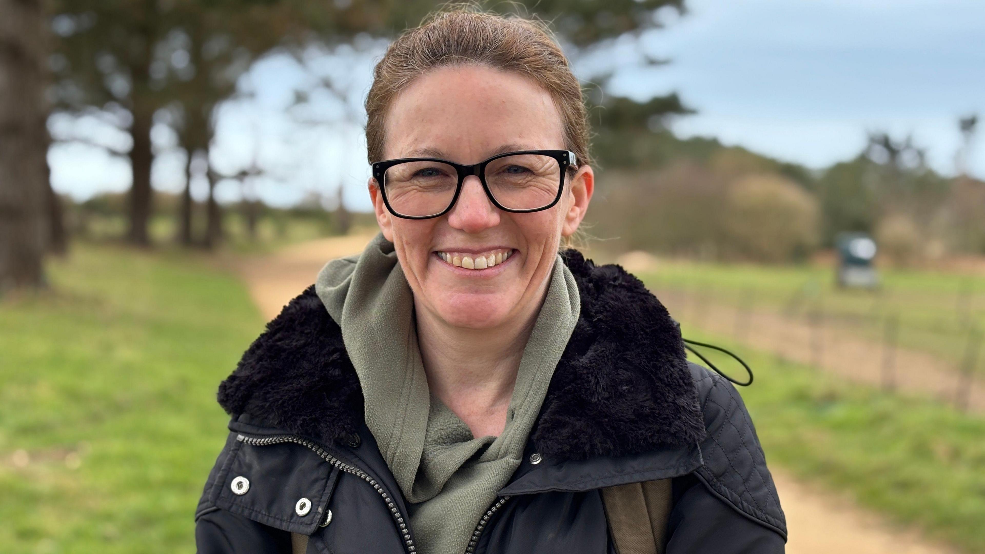 A woman with light brown hair is dressed in a green hooded jumper and a black winter coat. She has black rimmed glasses and is smiling while looking directly into the camera.