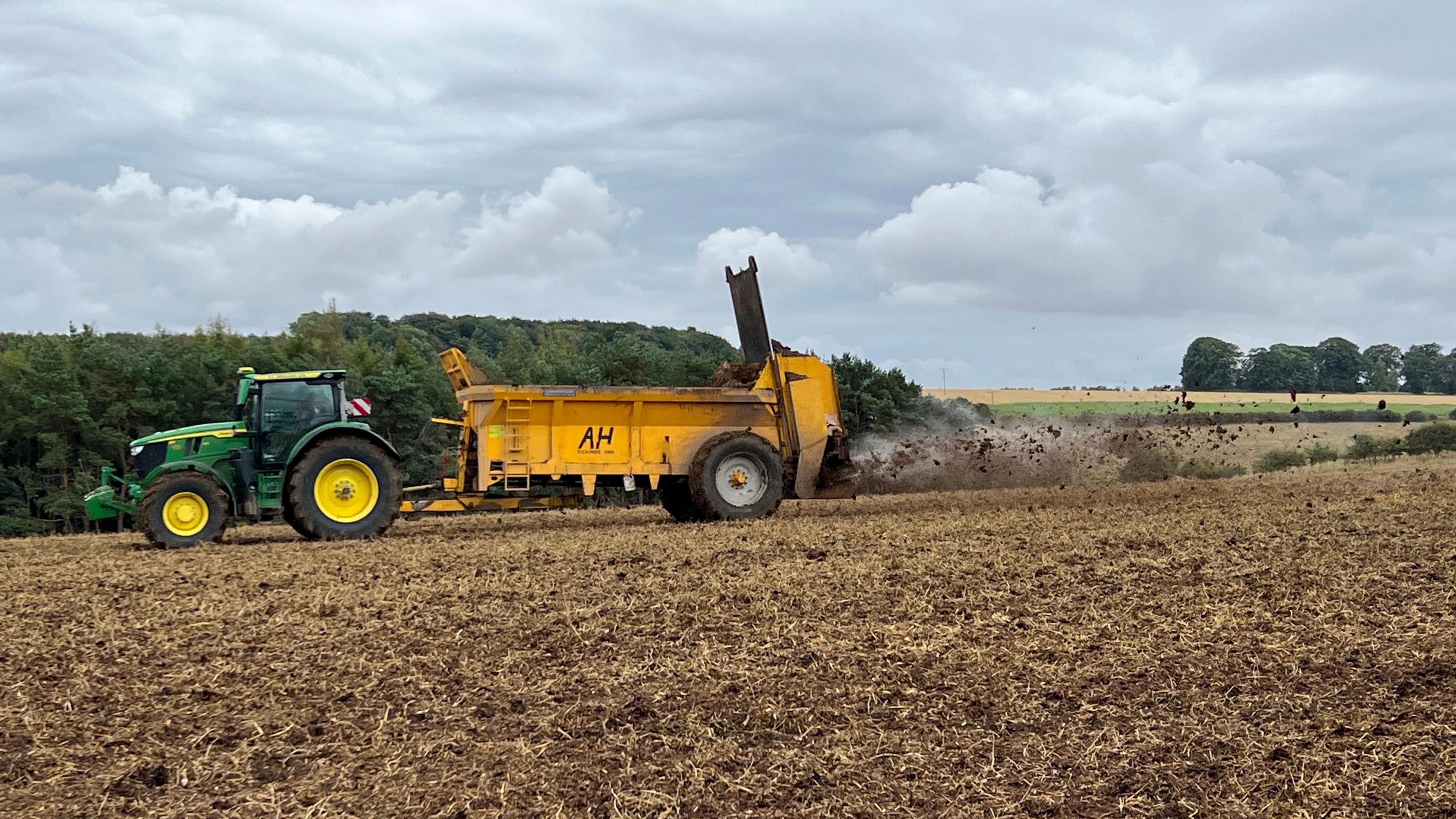 A green tractor is pulling a yellow trailer that is spreading muck on to a field of soil out the back of it.