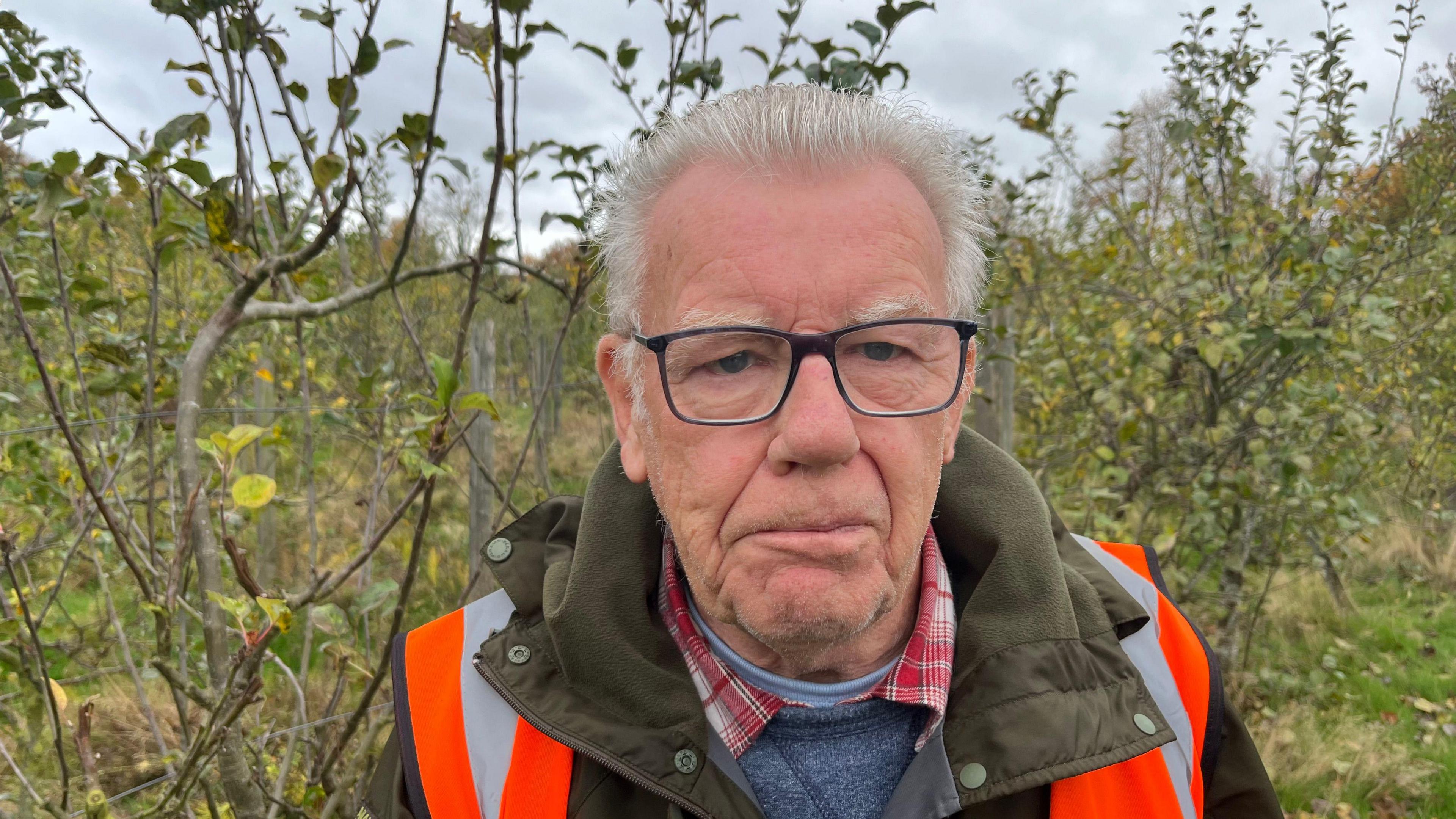 Roger Rawson,77, has short grey hair on top of his head and down the sides of his head, he has dark framed glasses on and a grey and orange high-viz tabard over a heavy dark green winter coat over a light blue woollen jumper over a red and white checked shirt over a blue t-shirt, behind him are trees which form an orchard.