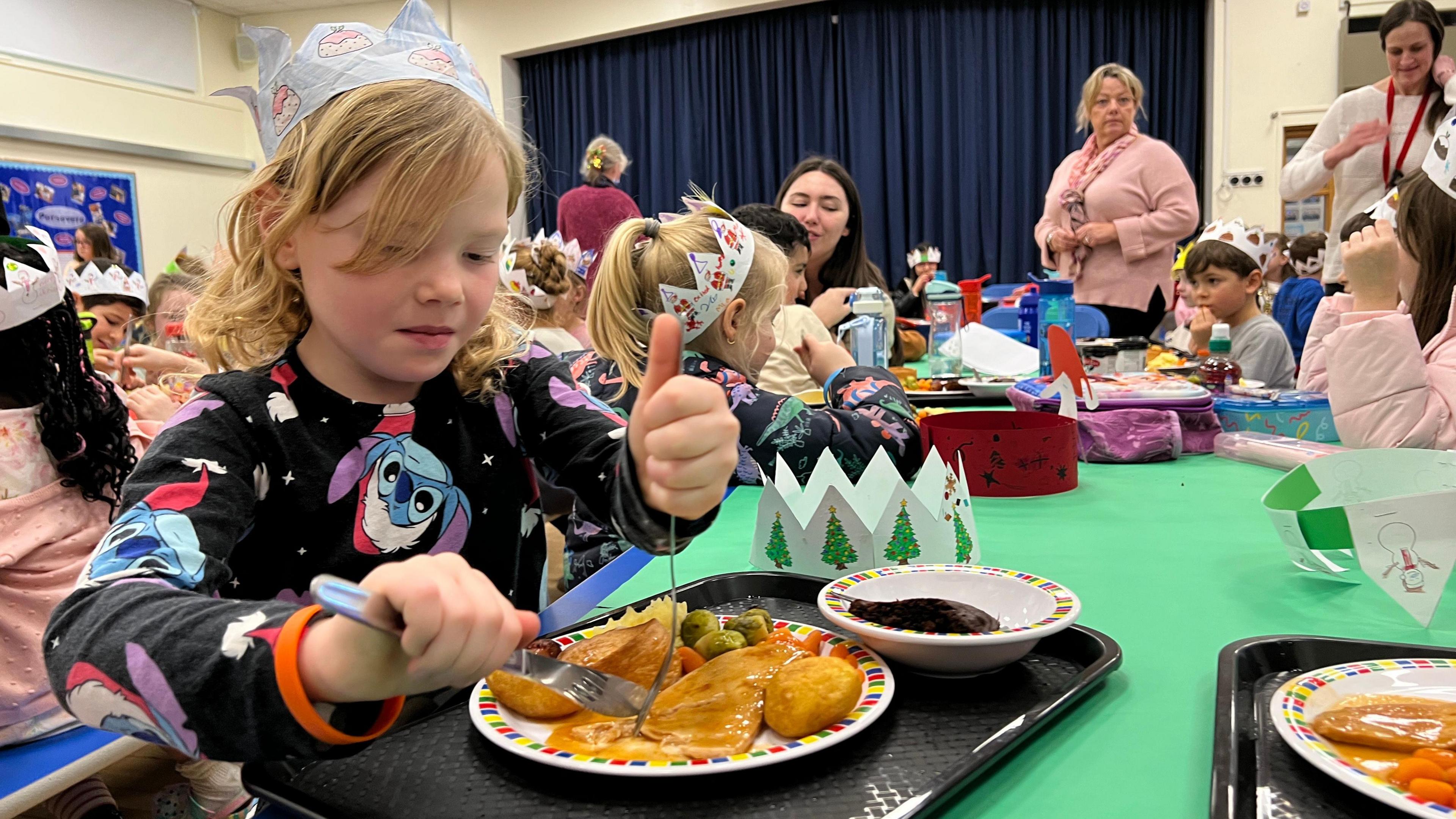 A young girl in cartoon pyjamas is holding a knife and fork as she cuts into some roast turkey on her plate which also has a roast Yorkshire pudding with Brussel sprouts. There's a bowl with a chocolate pudding next to her food. She's in a school hall with lots of other children sitting around her eating and she's also wearing a Christmas paper crown too.