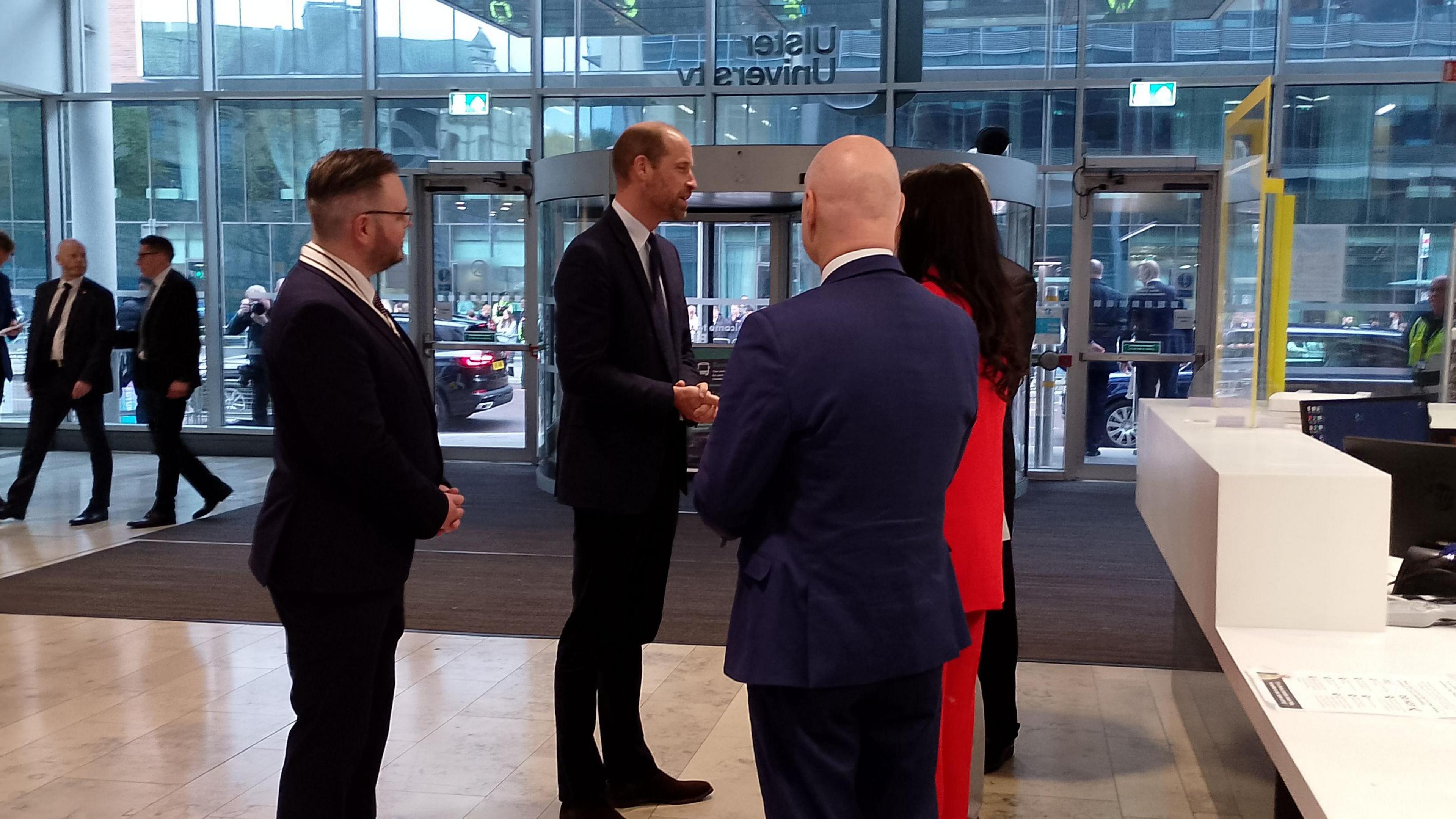 Prince William is standing with five unknown others. He is wearing a dark coloured suit. 