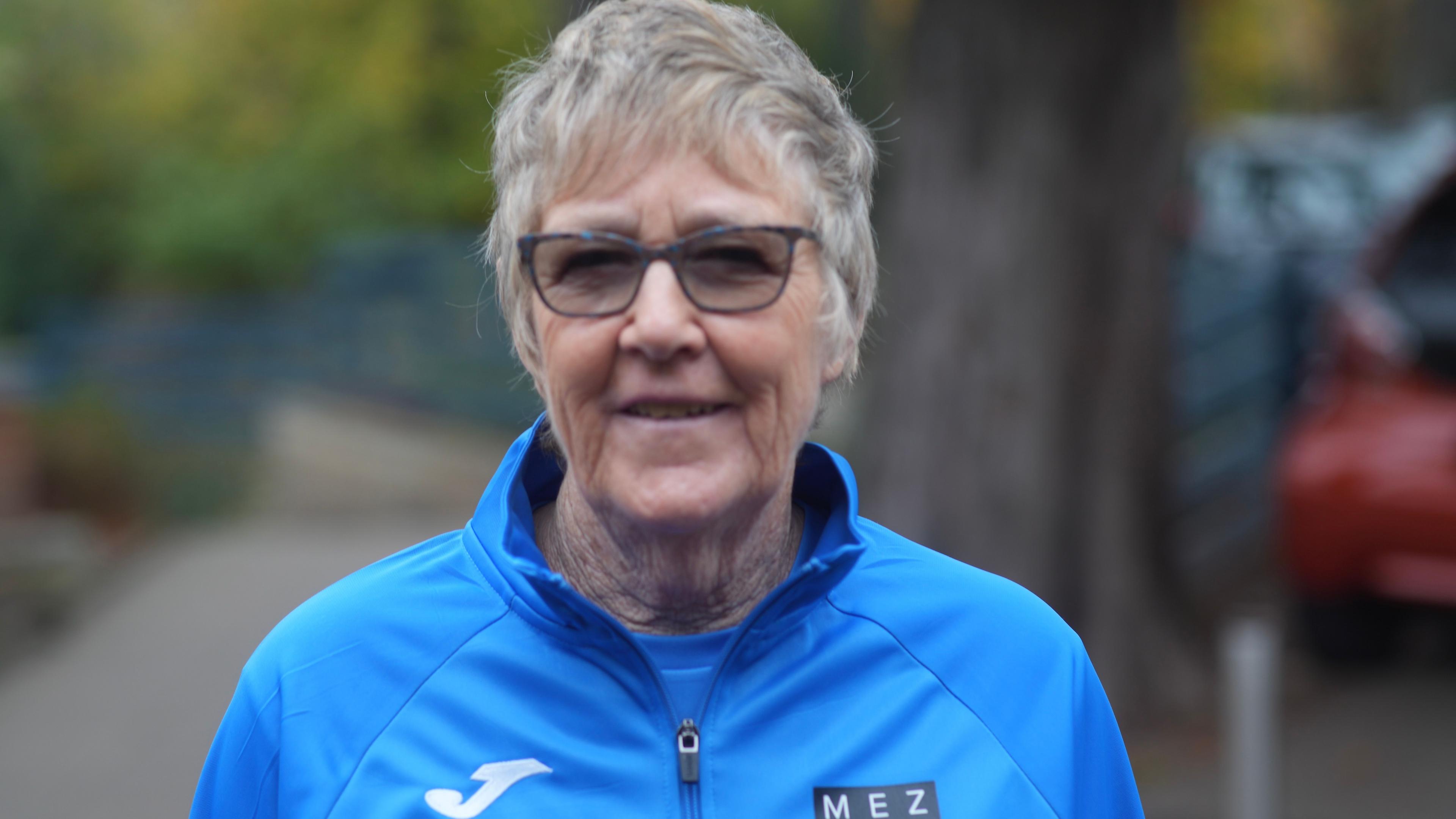 Penny Moor smiles at the camera. She is wearing dark glasses and she has short grey hair. She is wearing a blue jumper with a blue top underneath.