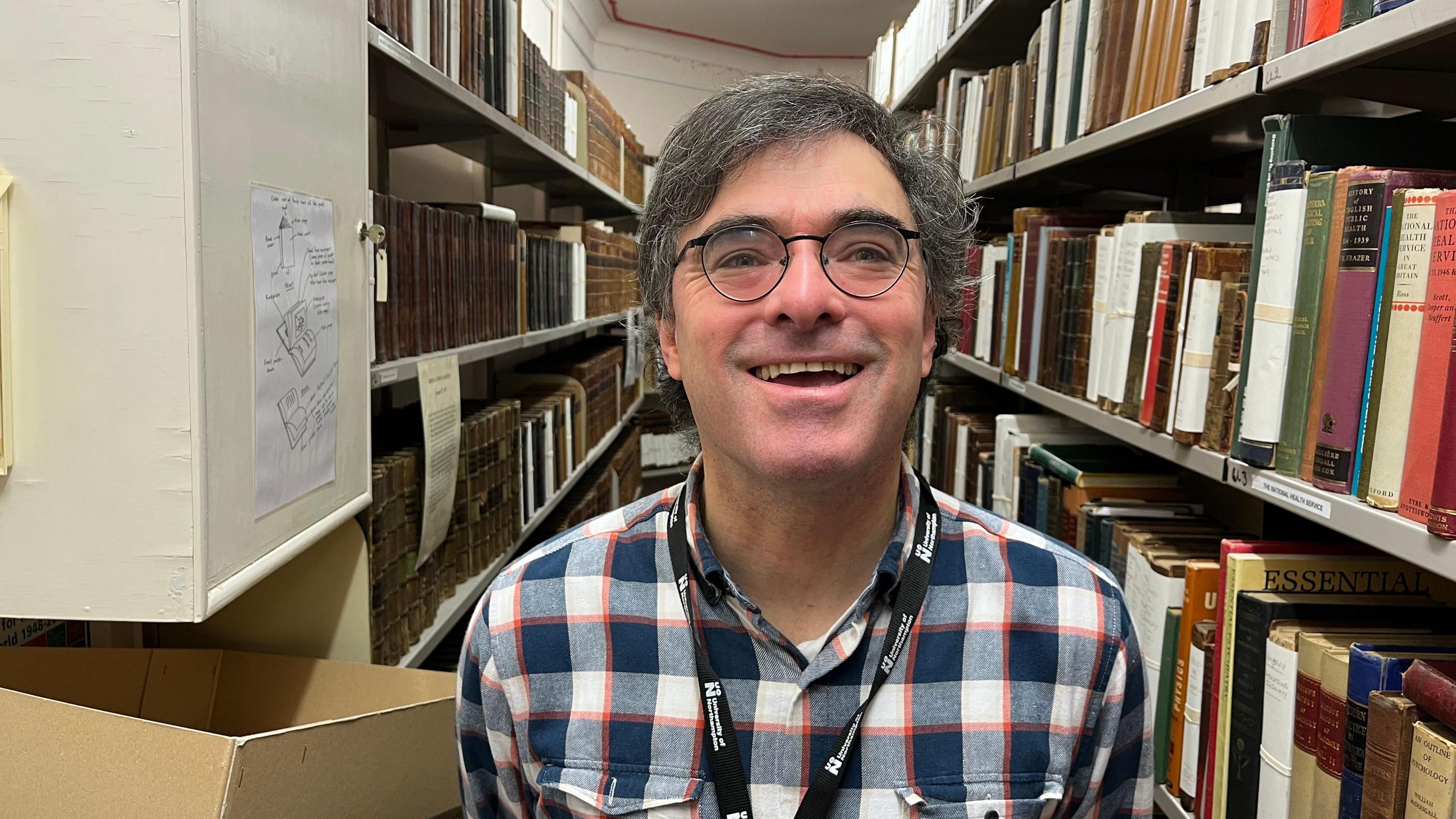 A smiling man in a check shirt and glasses stands in a corridor with lots of books on shelves on both sides of him.