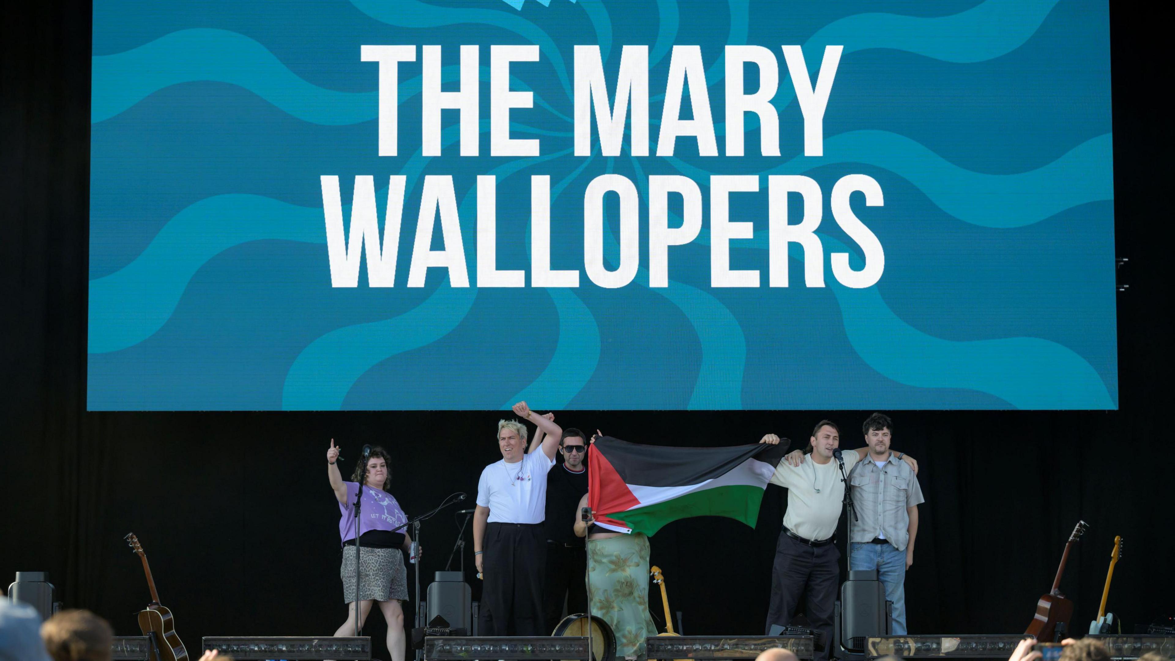 Multiple members of The Mary Wallopers stood on the Victorious Festival stage holding a Palestinian flag and with mouths open and arms raised appearing to chant. Behind them is a large screen with their band name on.