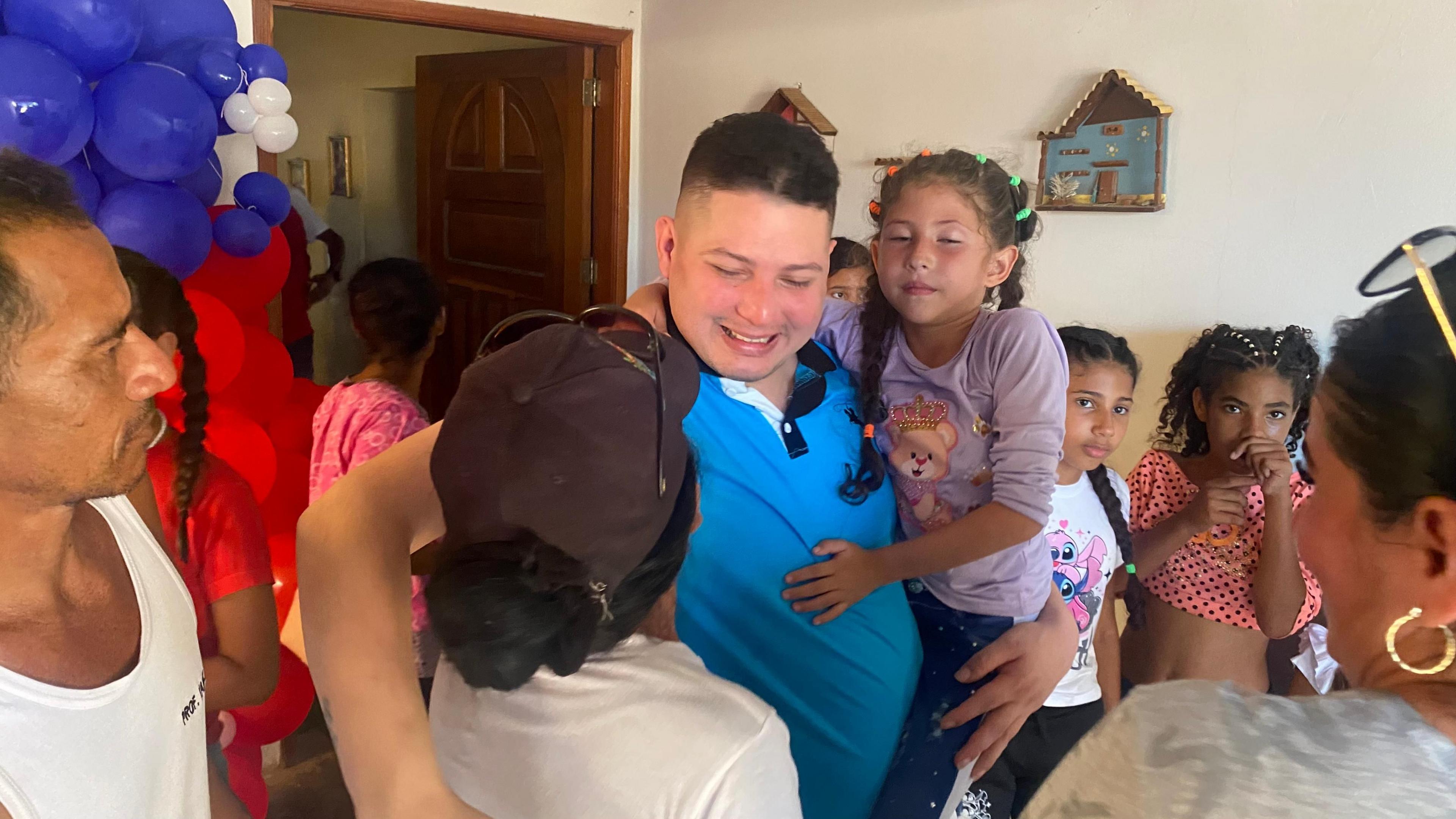 Andy Perozo, wearing a blue polo shirt and smiling, carries a young girl on his arm and embraces a woman. Other children look. In the background, balloons can be seen