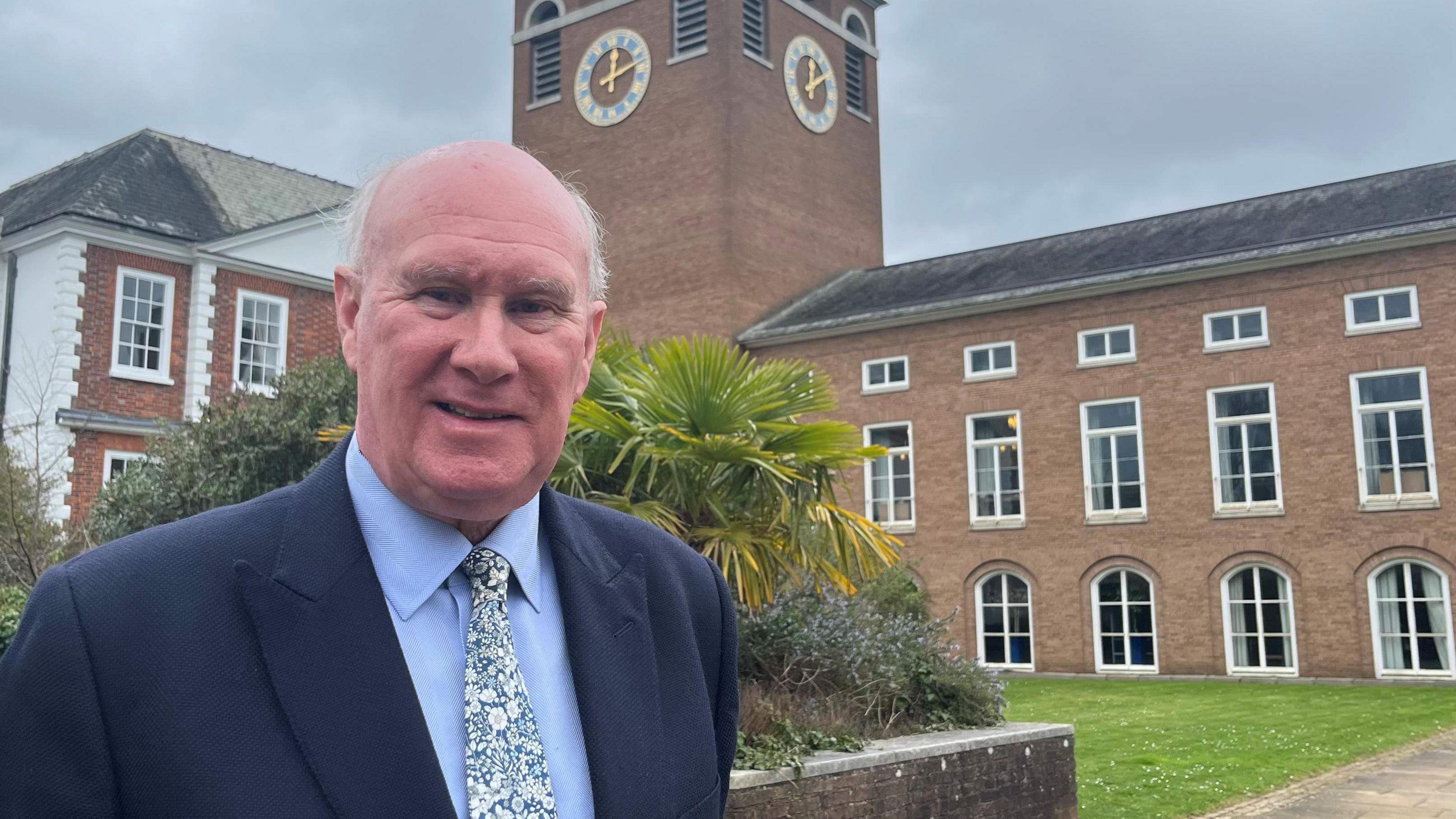 James McInnes, leader of Devon County Council, wearing a light blue shirt, a patterned tie and a navy blue jacket, standing in front of County Hall.