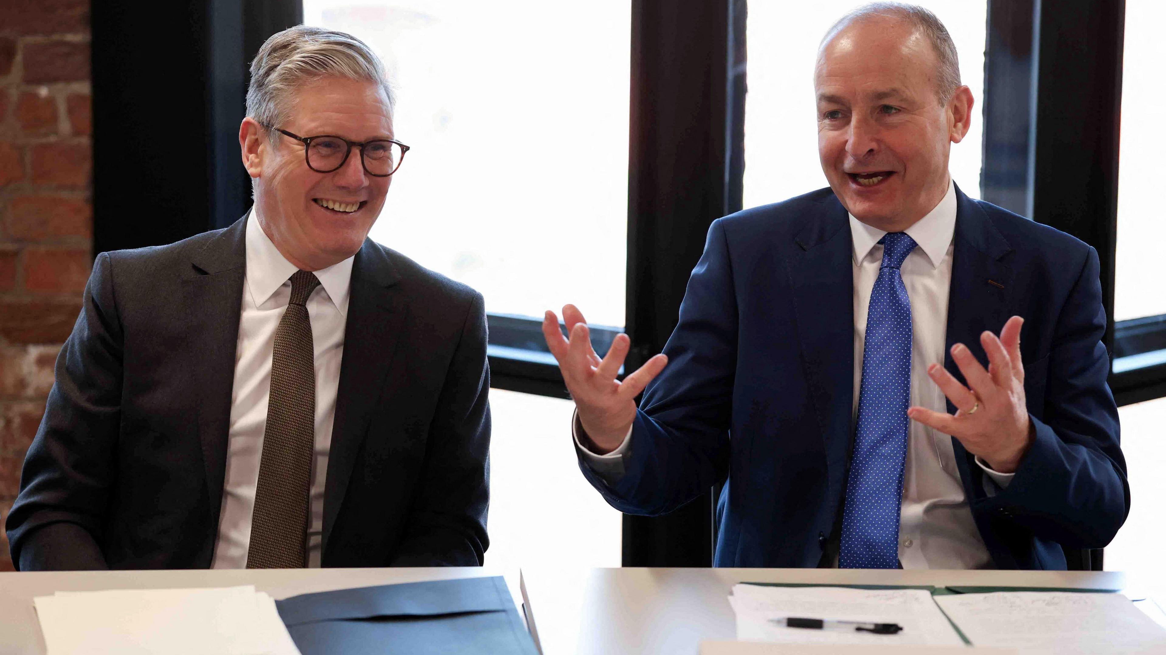 Keir Starmer (left) and Micheal Martin (right) sit at a white table. Both wear suits and there are papers in front of them. Both are smiling and Martin is gesturing with his hands. 