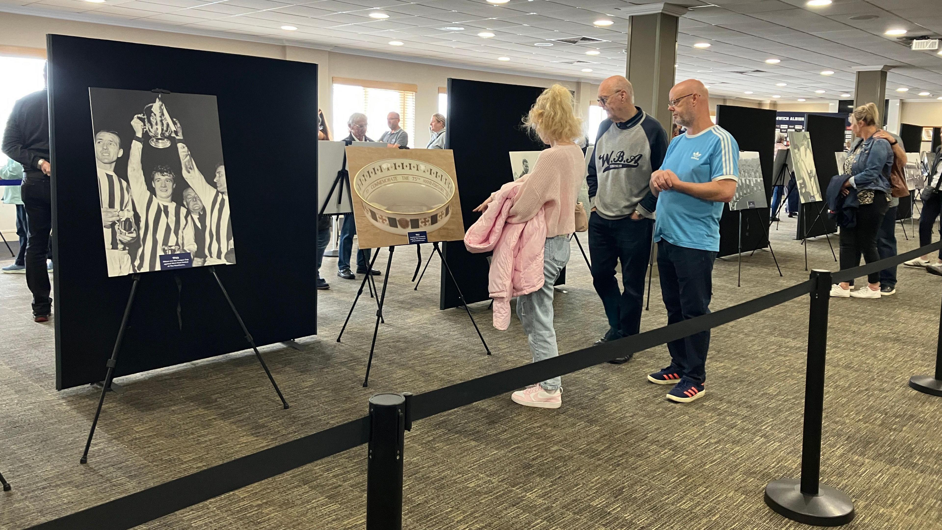 Fans gather round exhibits on display. Black boards have been set up with pictures of players and club memorabilia. A woman is standing in front of a picture with two men and other people are walking round the exhibition.
