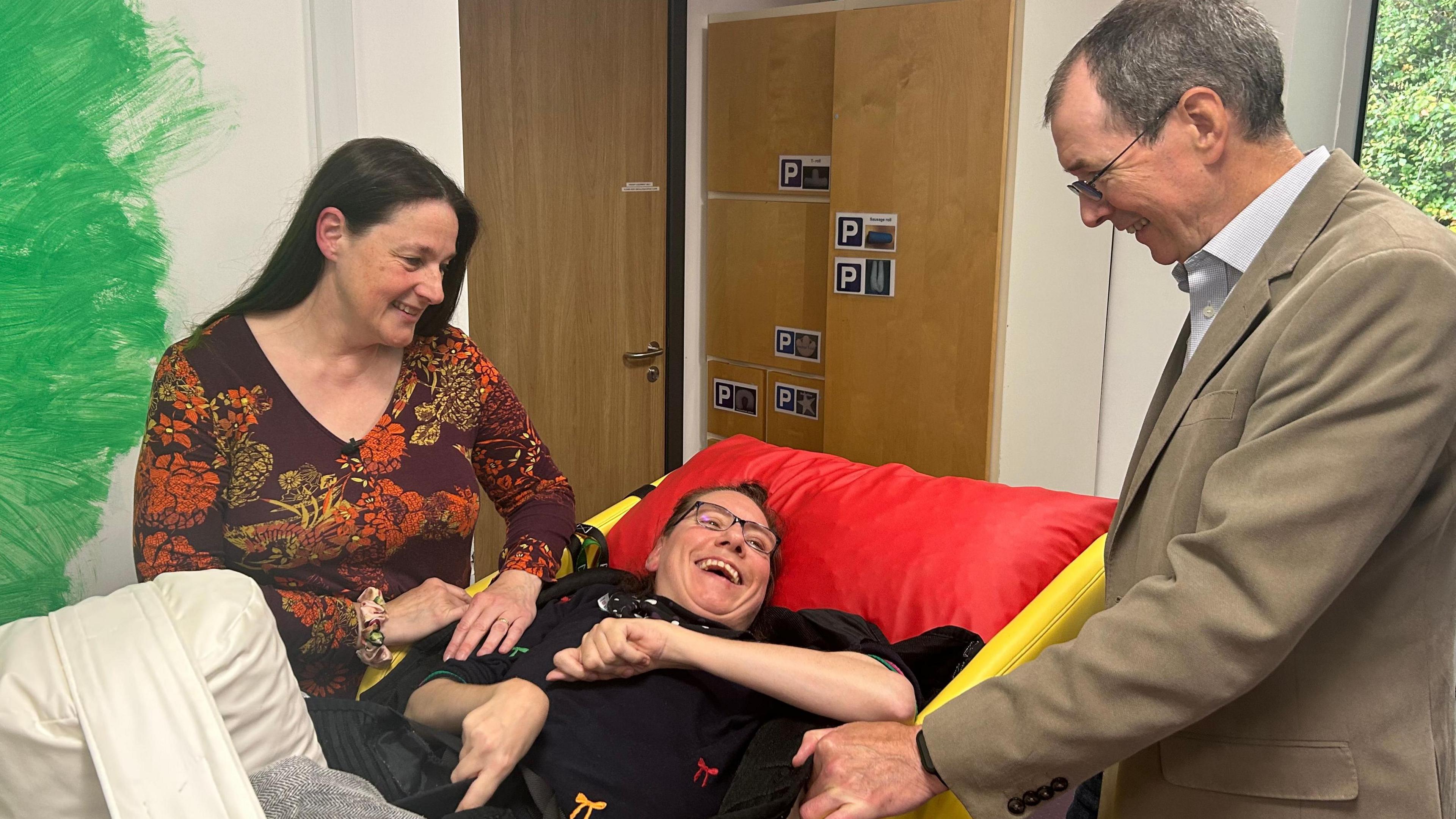 Tina Poole an occupational therapist, with long brown hair and a burgundy and orange floral top, looks down at Claire Dempsey, her patient who is smiling broadly at her dad, who stands on the other side of the bed.