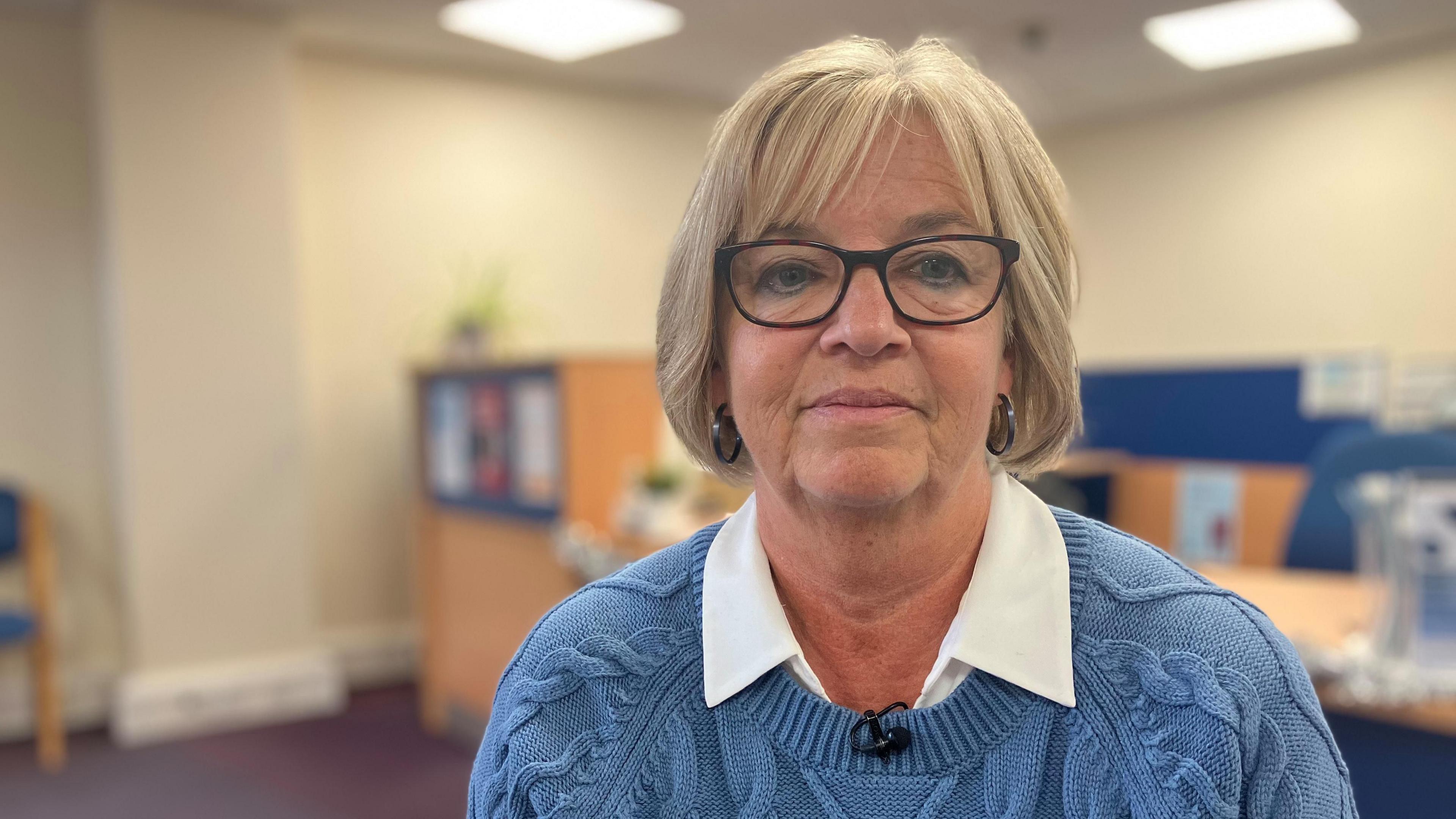 A woman with short blonde hair and dark-framed glasses and hooped earrings in a blue knitted jumper looks into the camera. 