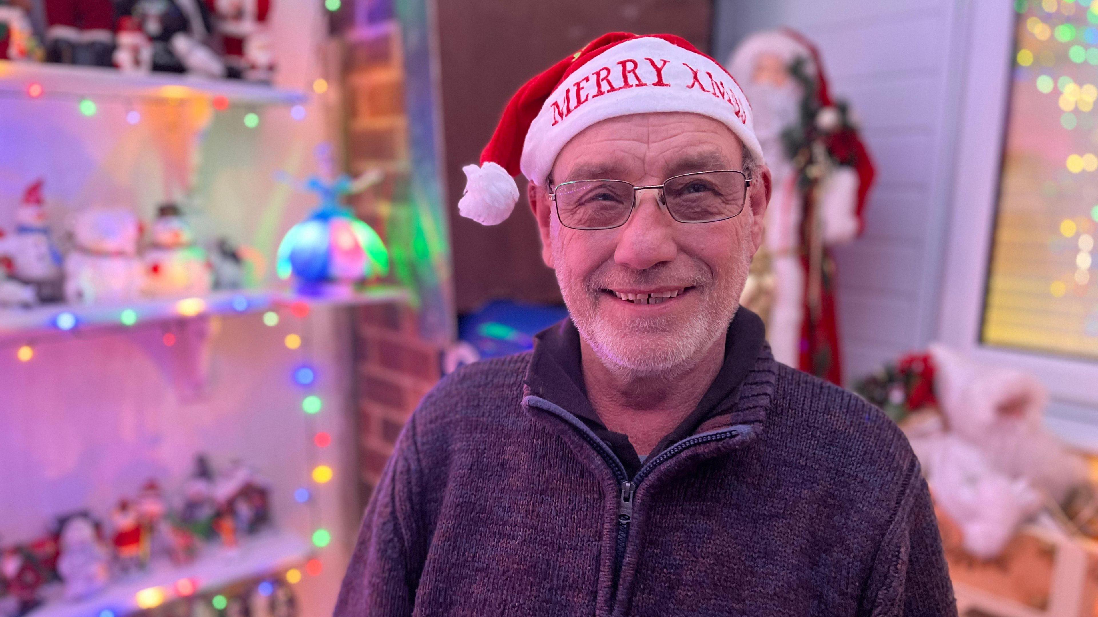 Frank Petchey wearing glasses smiles at the camera, while wearing a Christmas hat that reads "Merry Xmas". In the background are festive decorations, lights and a traditional-looking Father Christmas.