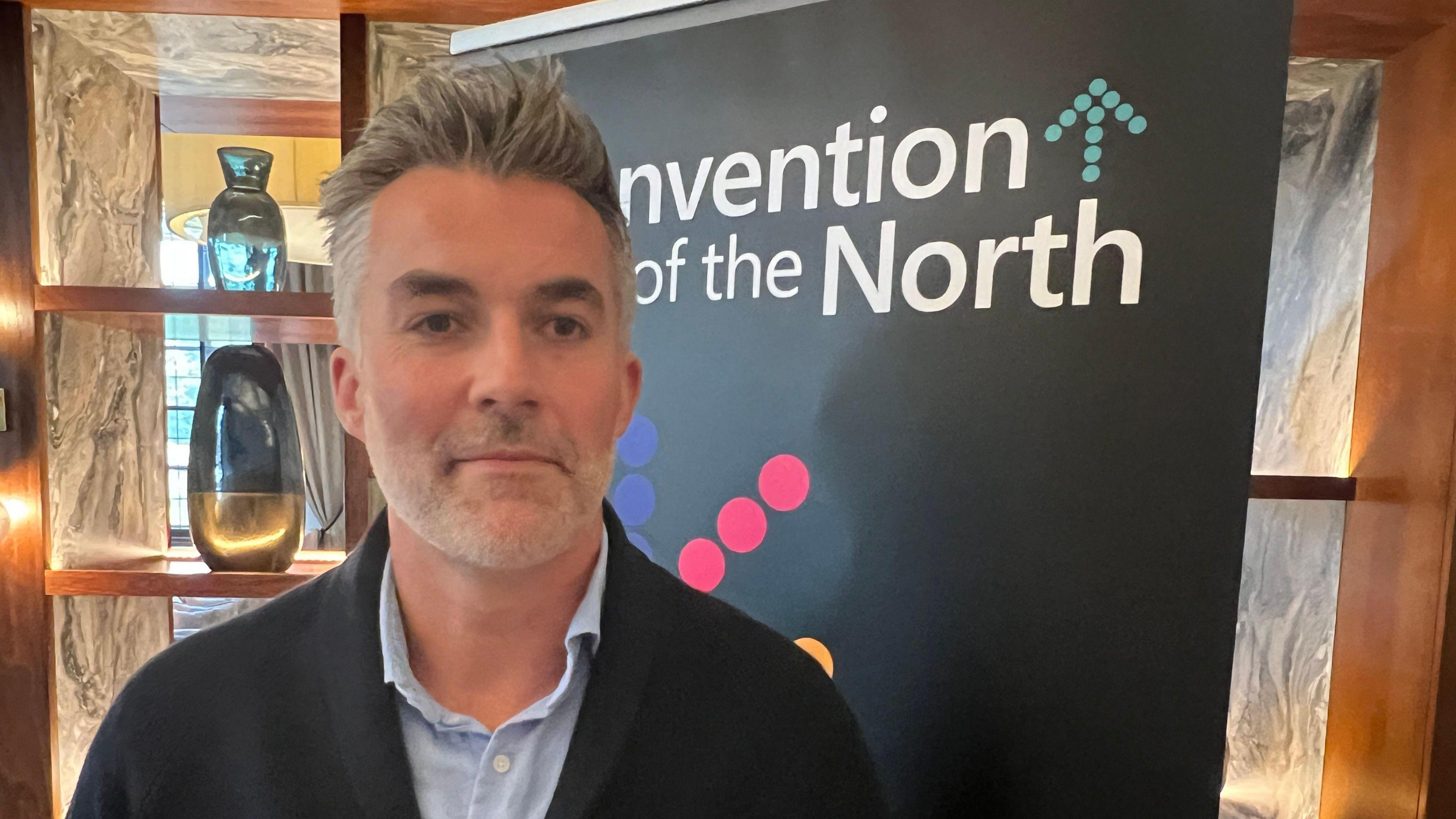 A photograph of Mr Skaith, who has medium-length grey hair styled in an upwards fashion. He is wearing a blue shirt and a black coat as he stands in front of a banner.