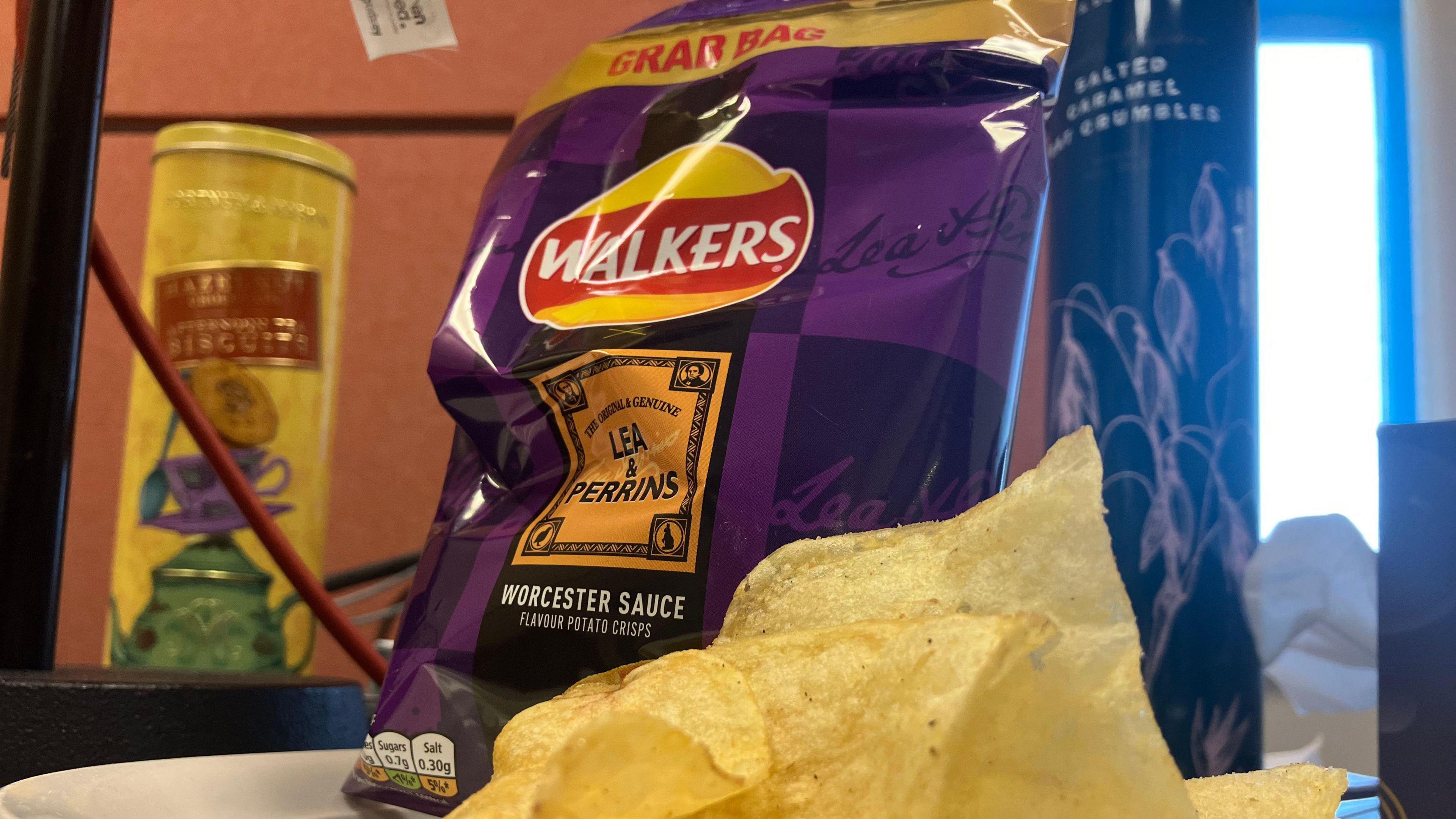 A white plate on a table containing a handful of crisps and a purple packet of Walkers Worcester sauce crisps. A wire and tubes of other food stuffs can be seen behind.