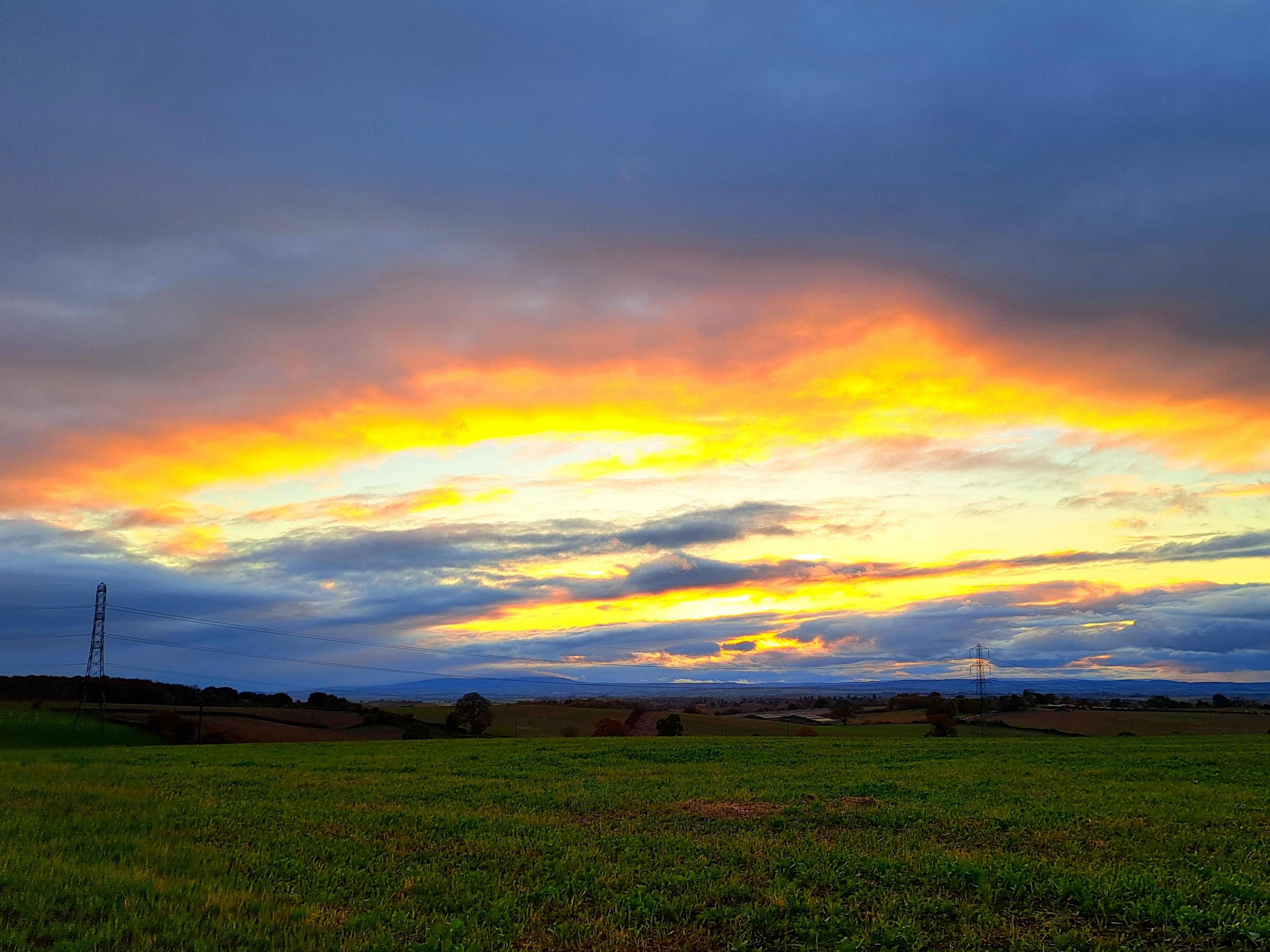 A view across fields with a string of pylons stretching into the distance, and a sky dominated by a setting sun shining through clouds