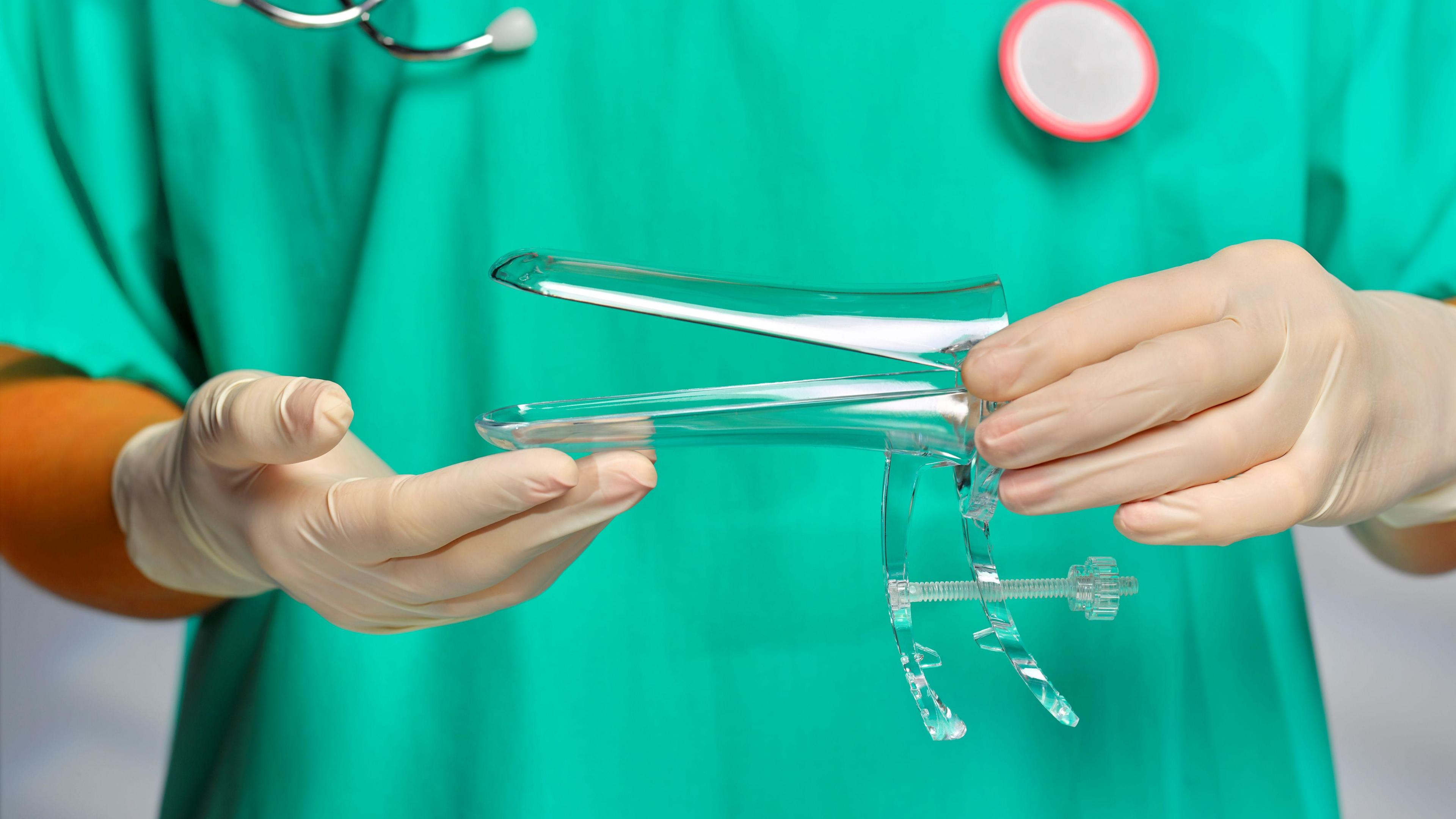 Gynecologist holding vaginal speculum. The gynecologists uniform is green and they are wearing disposable gloves. A stethoscope is around their necks