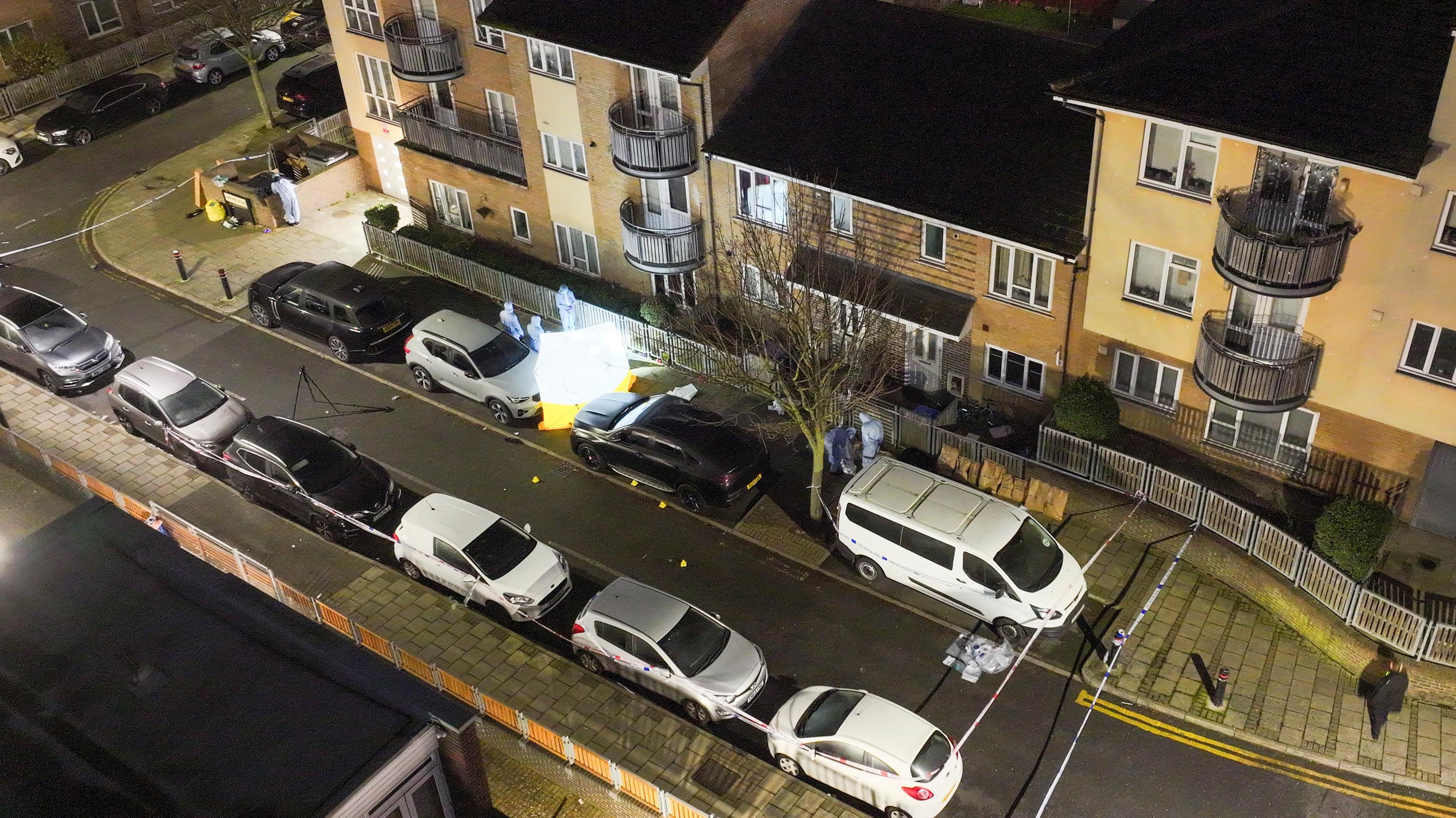 Aerial view of a cordoned-off crime scene with forensic investigators, evidence markers, and a tent set up on a residential street at night.