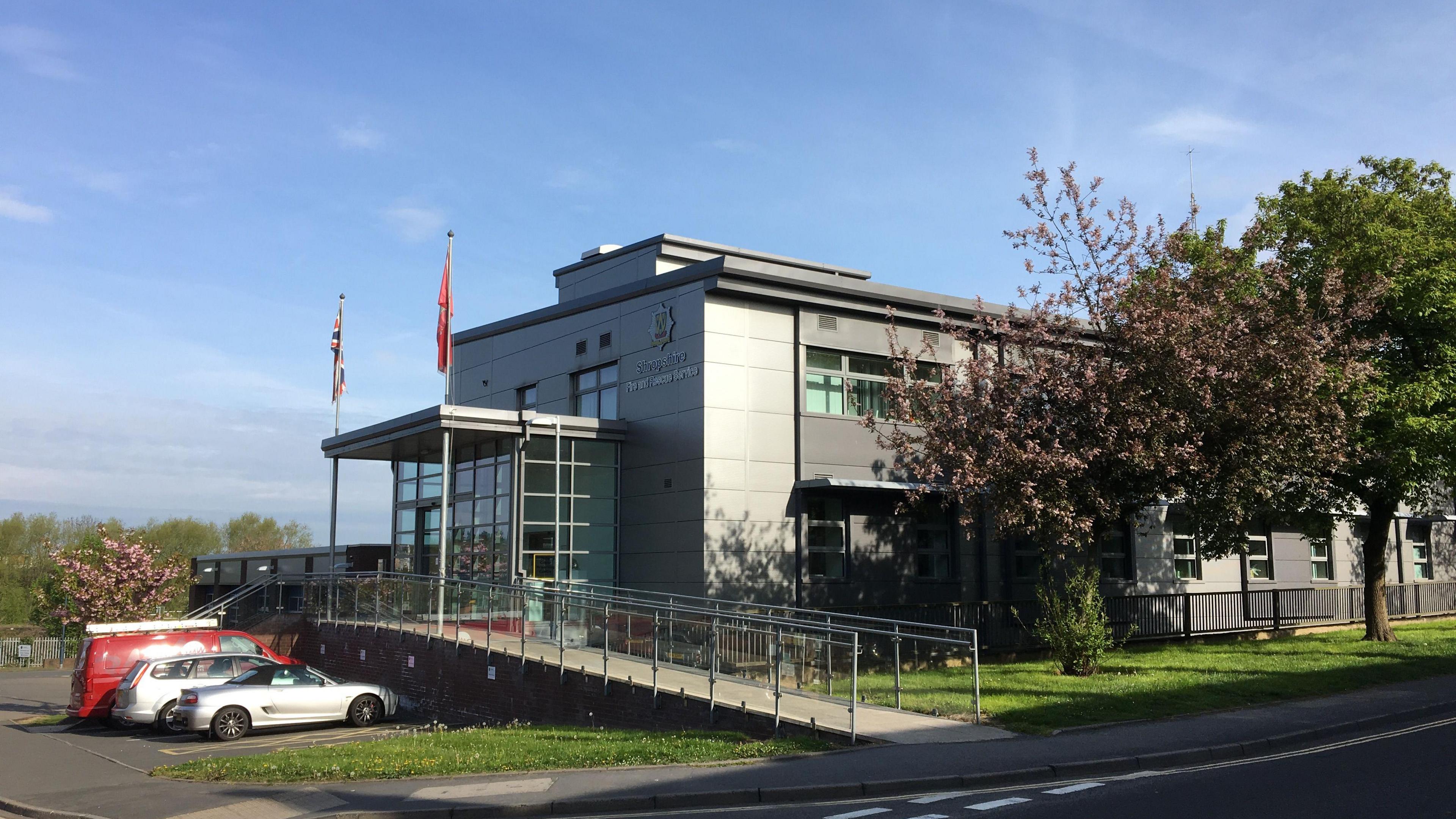 Shropshire fire and rescue service headquarters. There is a road adjacent to it. The building is grey and has two flags above the entrance. There is a ramp with glass panelling either side of it. There are trees on an area of grass to its right.