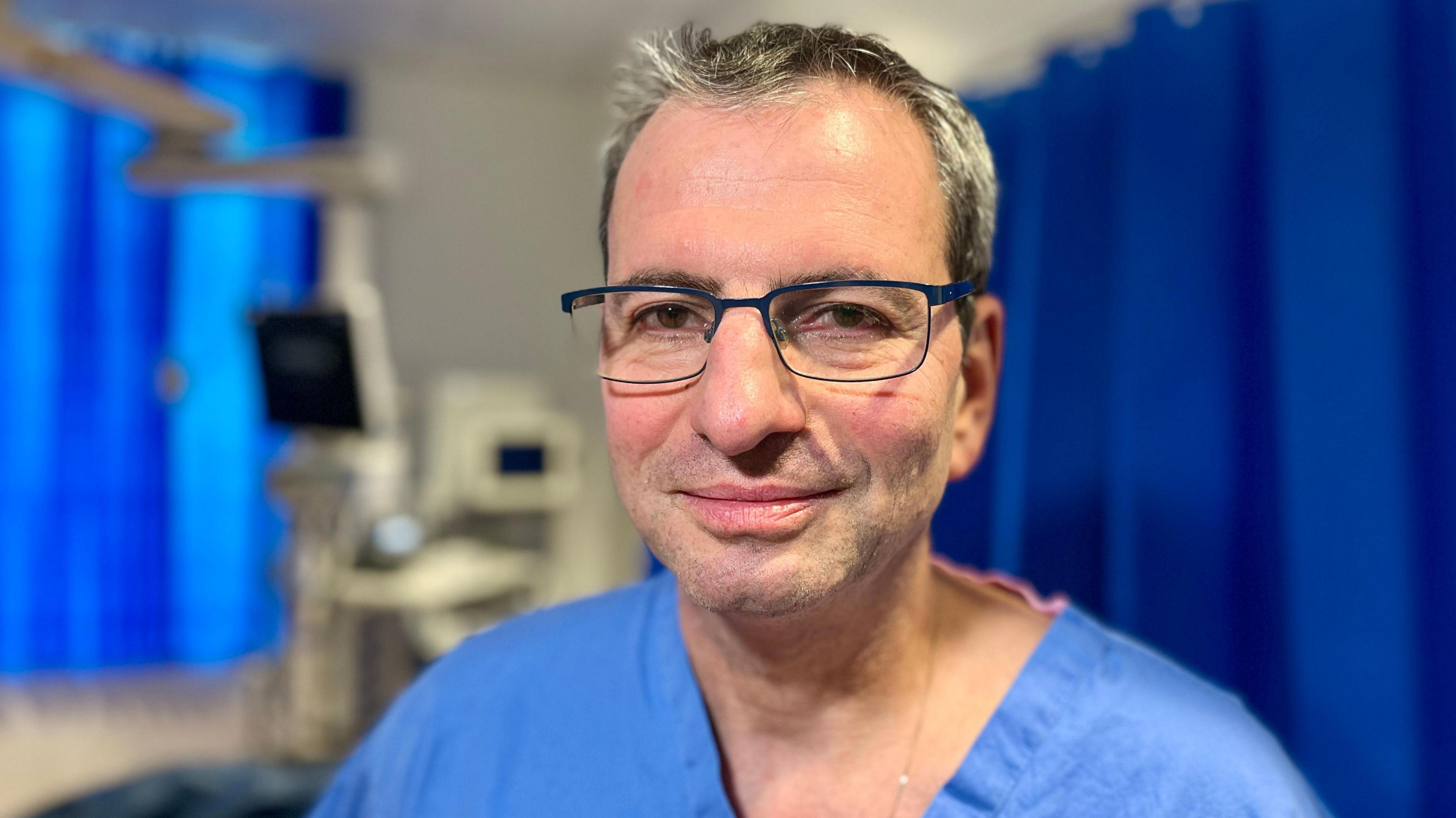 Surgeon Marius Berman standing in the critical care department at Royal Papworth Hospital in Cambridgeshire. There are blue curtains either side of him and some monitoring computers in the background which are blurred. he is wearing blue scrubs and has metal rimmed glasses on. He is looking directly at the camera with short greying hair.