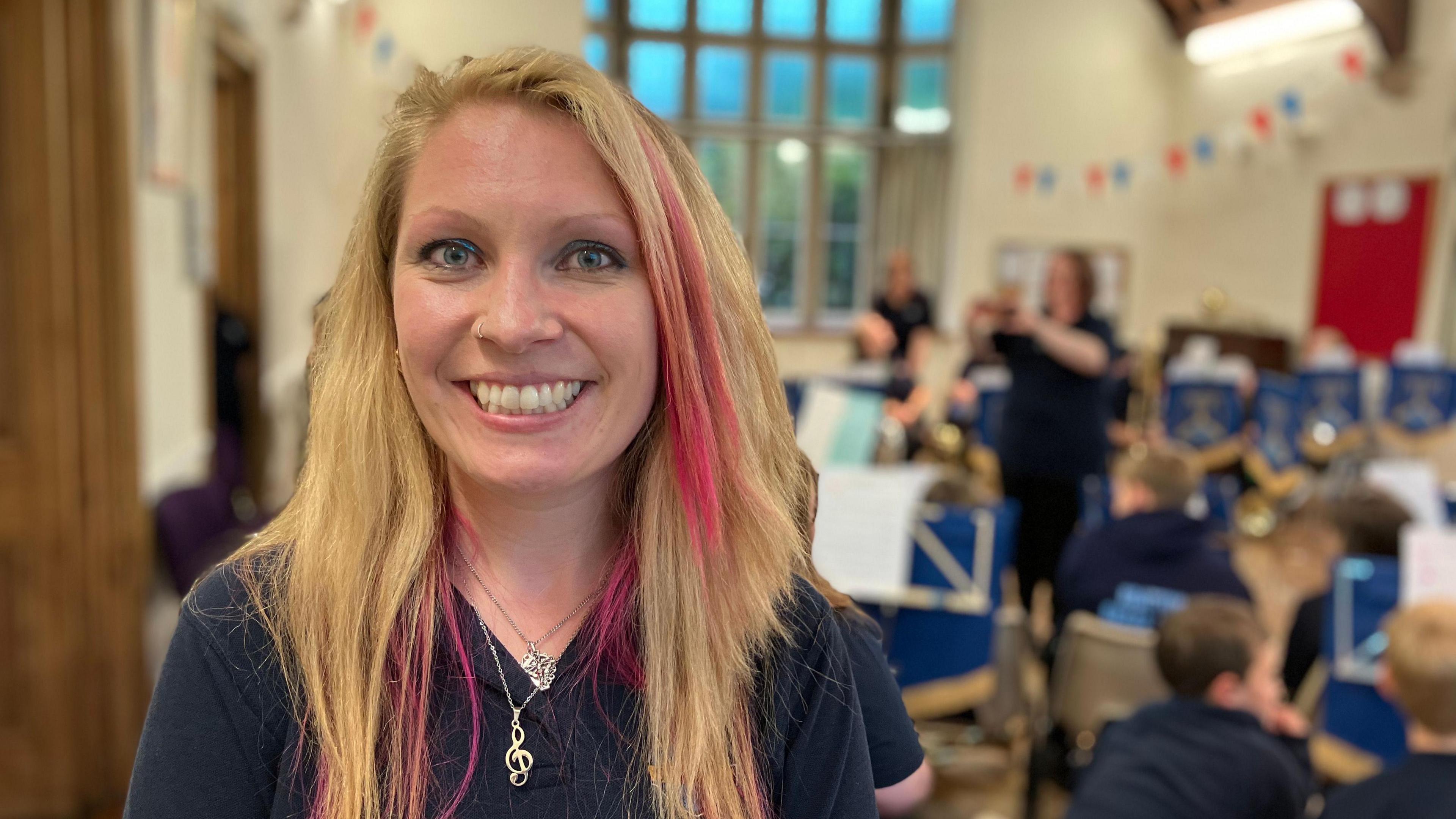 Lydia Grace-Hills standing in a room with her students behind her. She has blond hair with pink streaks and is wearing a dark shirt. She is smiling and looking at the camera.