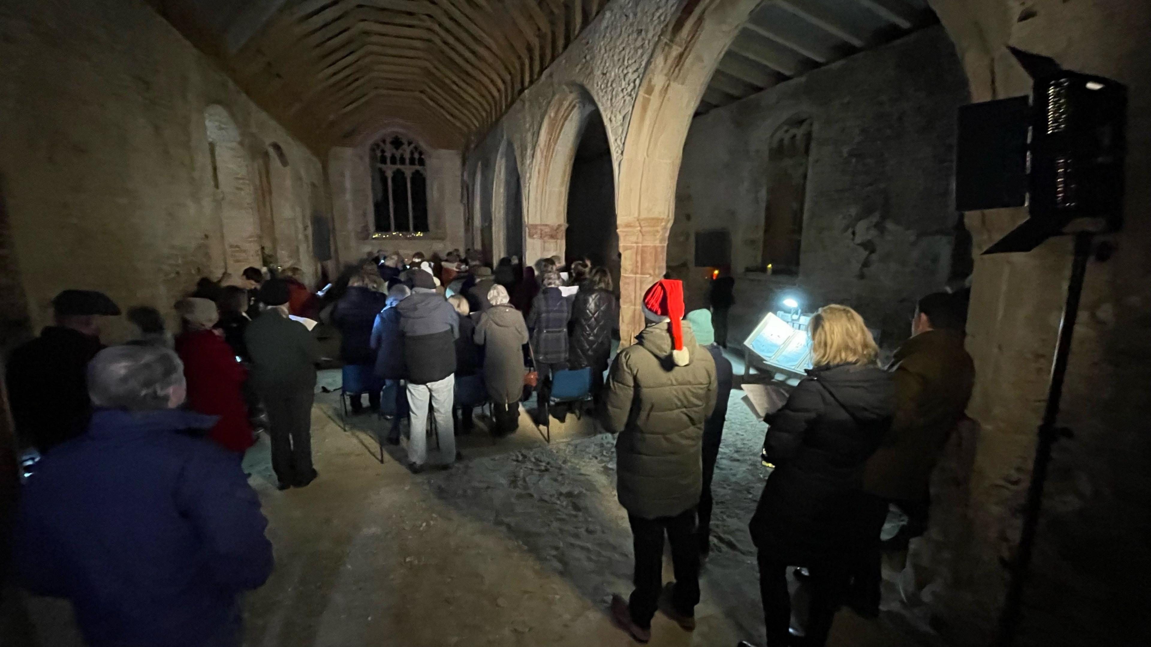 Congregation at a church service. They are standing in the nave which has bare floors, no pews and it is quite dark with theatre-type light on top of a pole on the right of the picture. One man is wearing a santa hat.