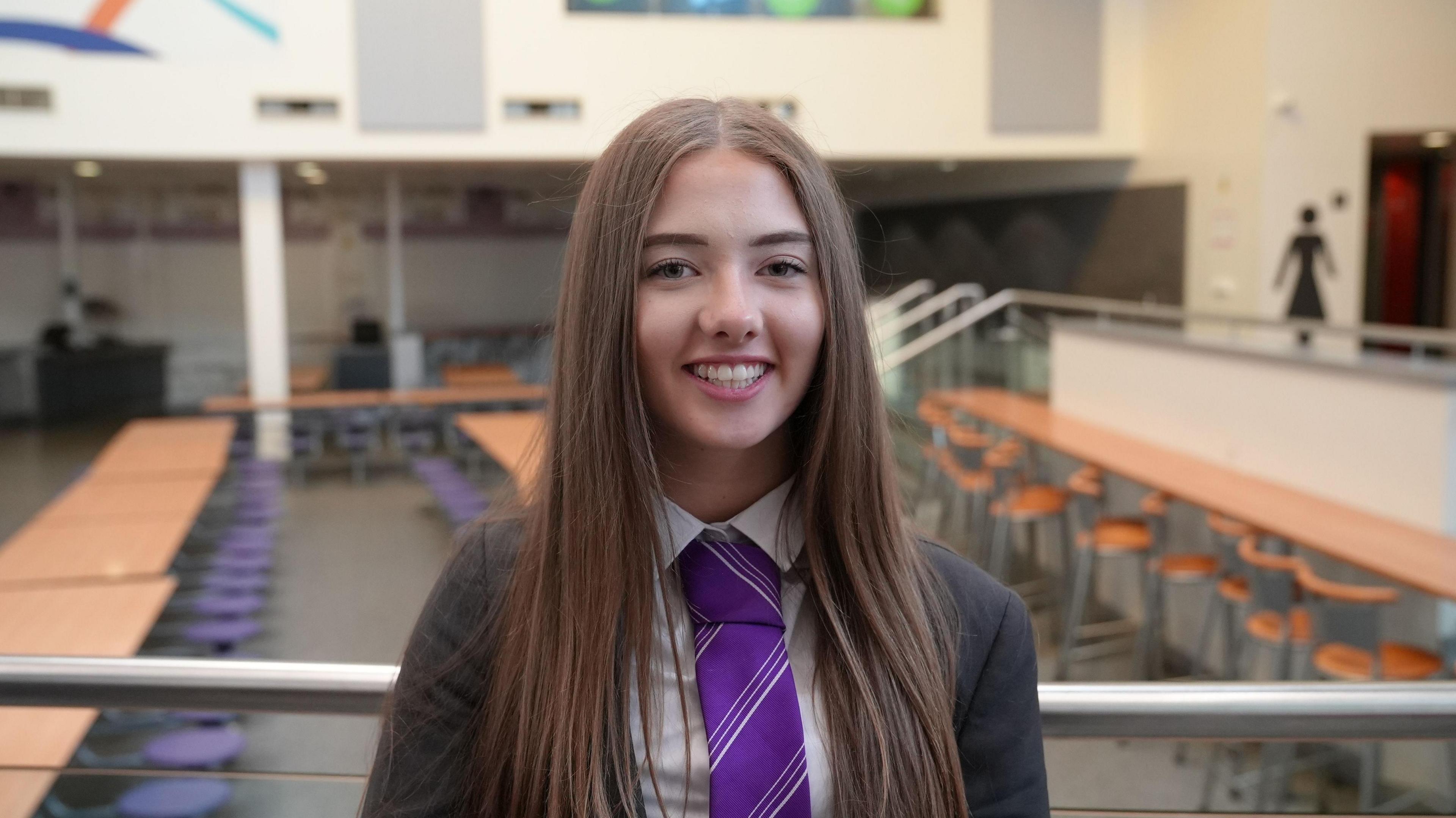 A teenager with long straight dark hair smiles at the camera. She is wearing a dark school blazer and a purple tie.