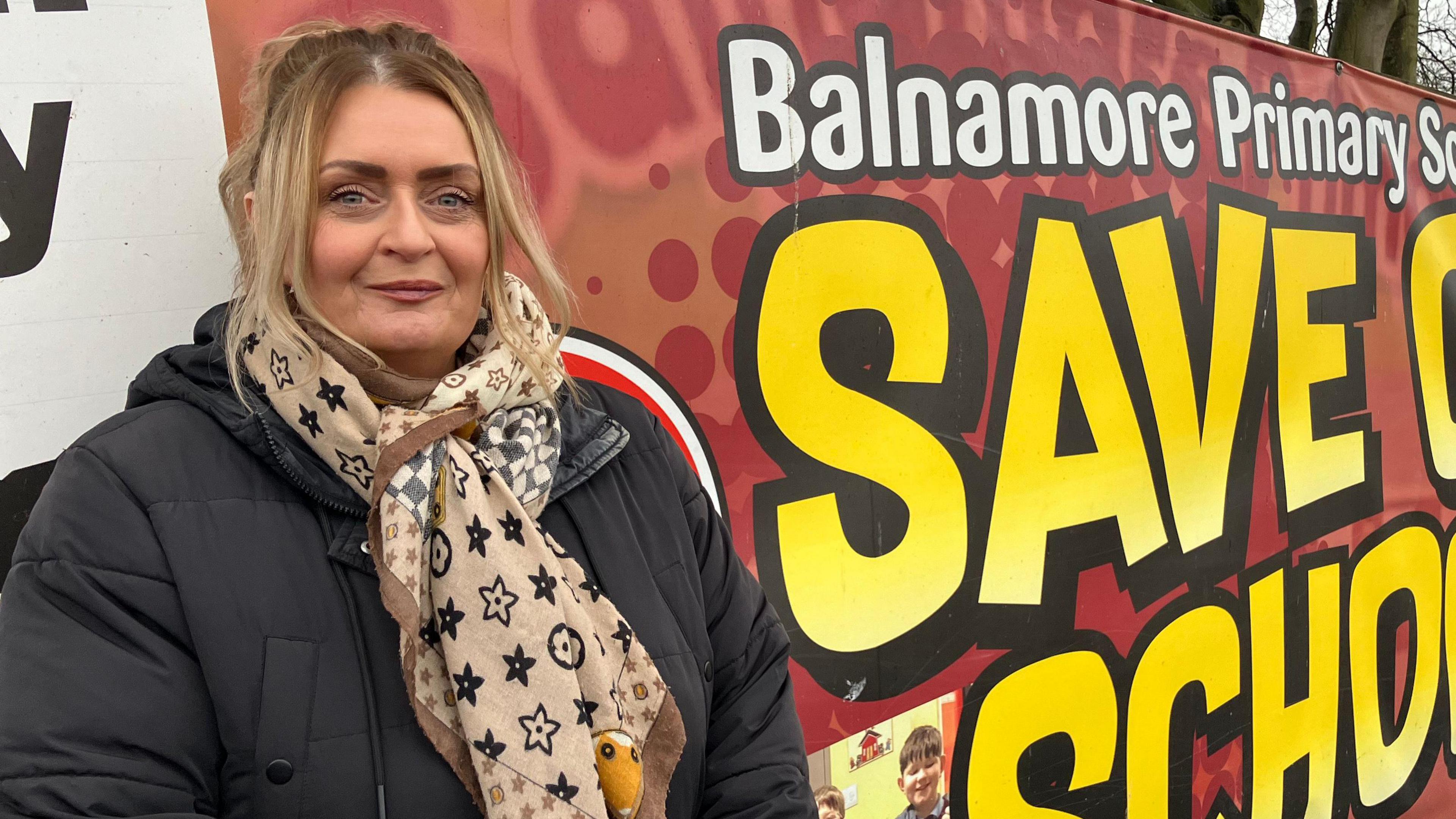 Joanne Skinner is looking into the camera. She's wearing a brown scarf with star patterns on it and a padded black coat. A large sign saying save our school is behind her.