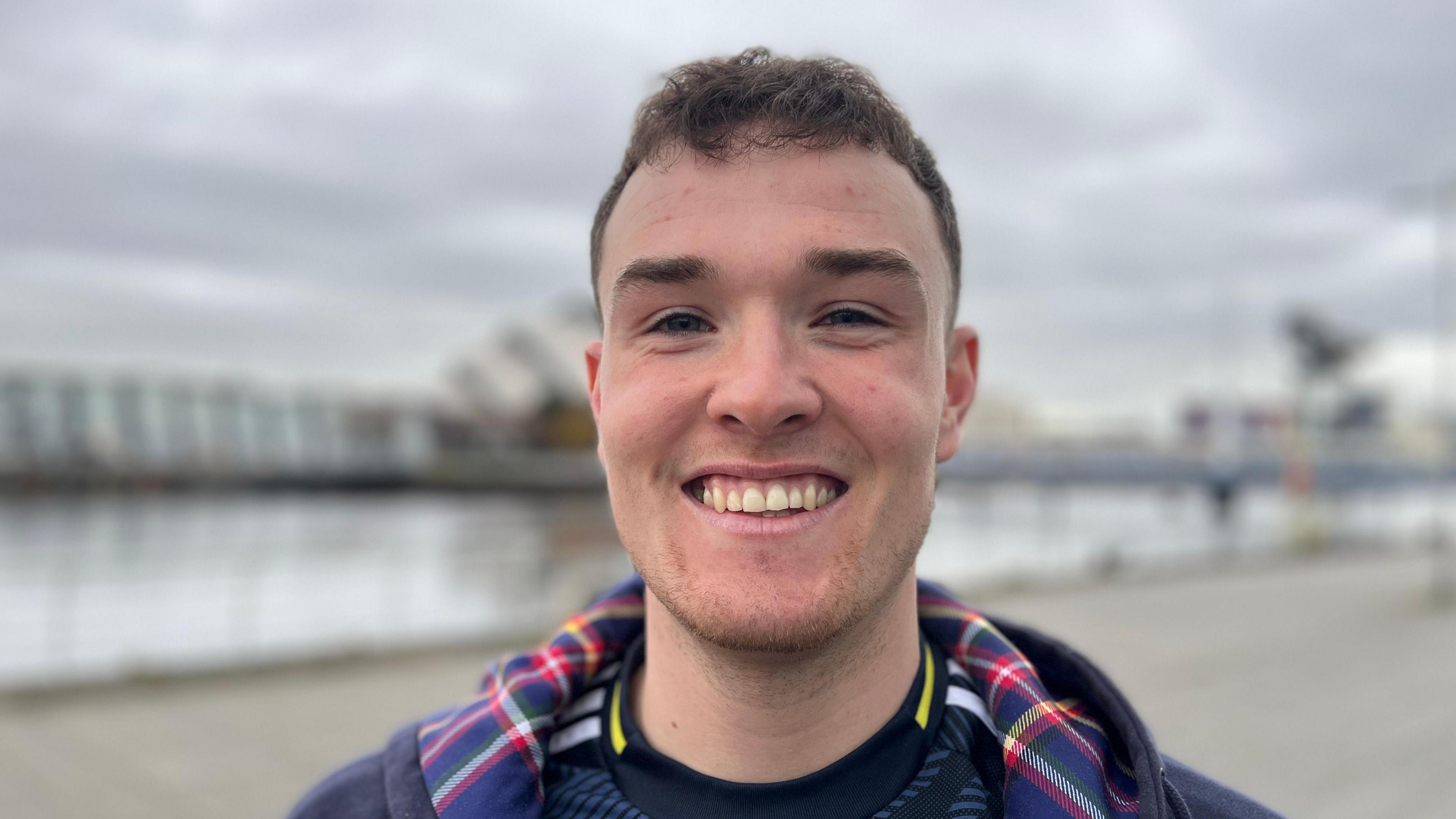 Craig Ferguson smiling and looking right at the camera. He is standing in front of the quayside in Glasgow which is blurred in the background. He is wearing a hooded top with a tartan lining in the hood and a dark blue Scotland football shirt with a blue and yellow collar and white lines on the sleeves.