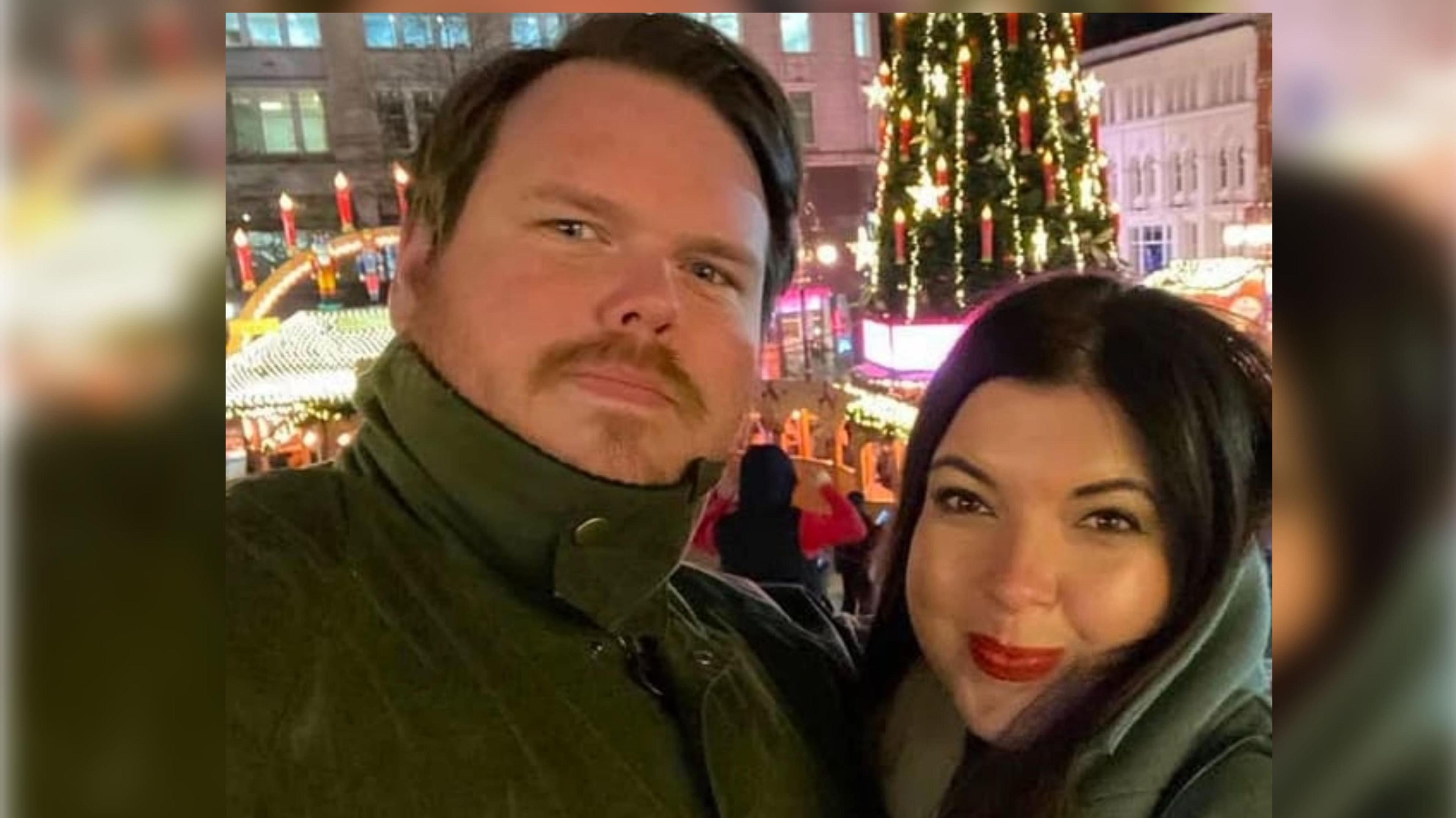 Steven (left) and Hannah Haggerty are taking a selfie. There is a lit up Christmas tree in the background. Steven Haggerty has brown hair and a brown moustache. He wears a green anorak buttoned up to his chin. Hannah Haggerty has black hair and is smiling with a closed mouth. She is wearing a green coat.