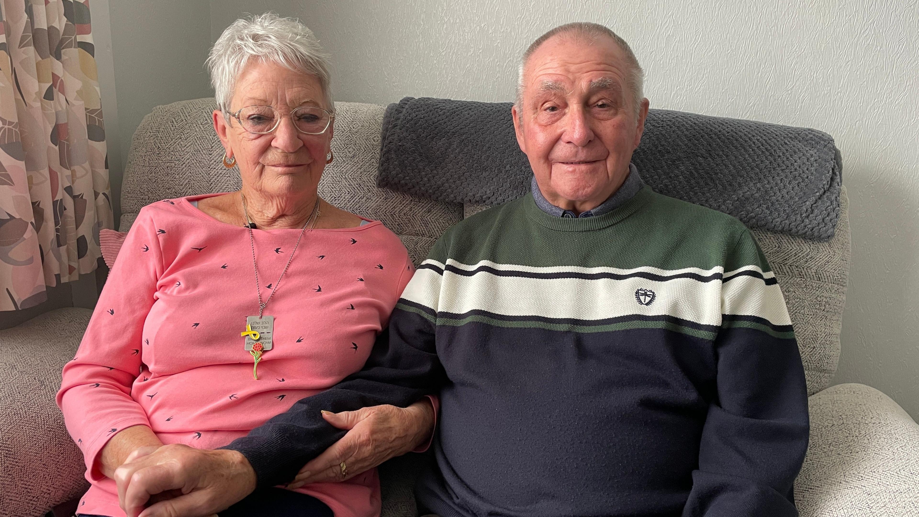 An older lady with short white hair, glass and a pink top with spots sits next to her husband on a sofa. She is holding his arm. The man has close cropped hair, and is wearing a green, cream and navy striped jumper. 