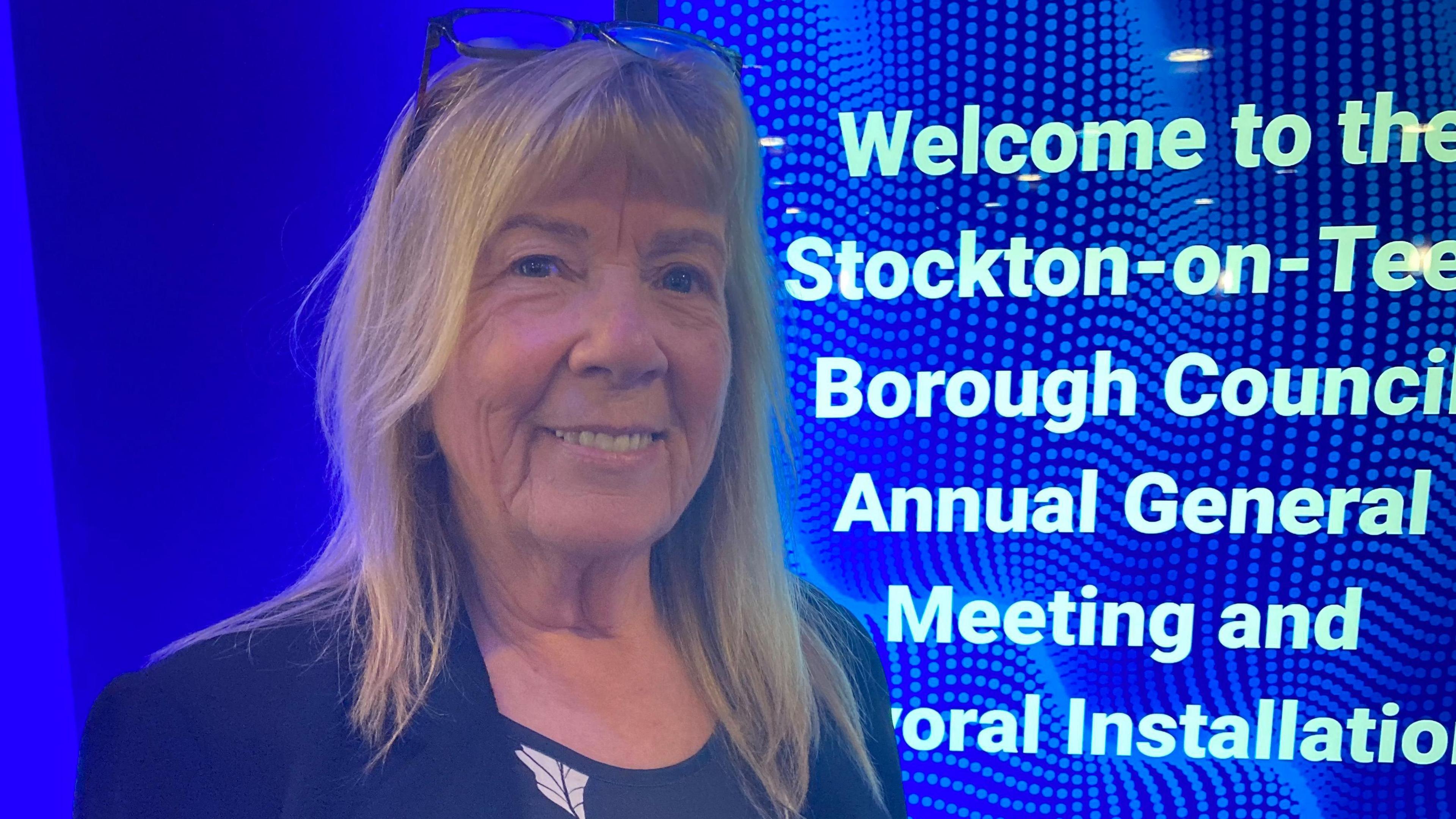 Head-and-shoulders image of Councillor Norma Stephenson. She has shoulder-length blonde hair with a fringe and is smiling into the camera. She is wearing a dark blazer and top, and standing in front of a sign welcoming people to a Stockton Council meeting.