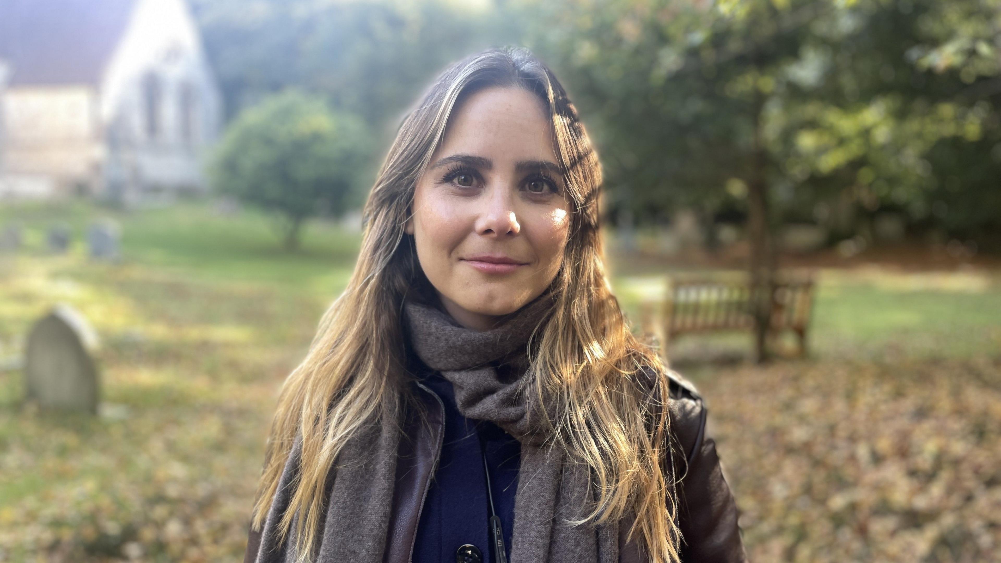 Rachel Hyde, who has long, blonde/light brown hair looks directly at the camera. She is standing in a churchyard. There are leaves on the ground the church is in the background. She is wearing a grey scarf.