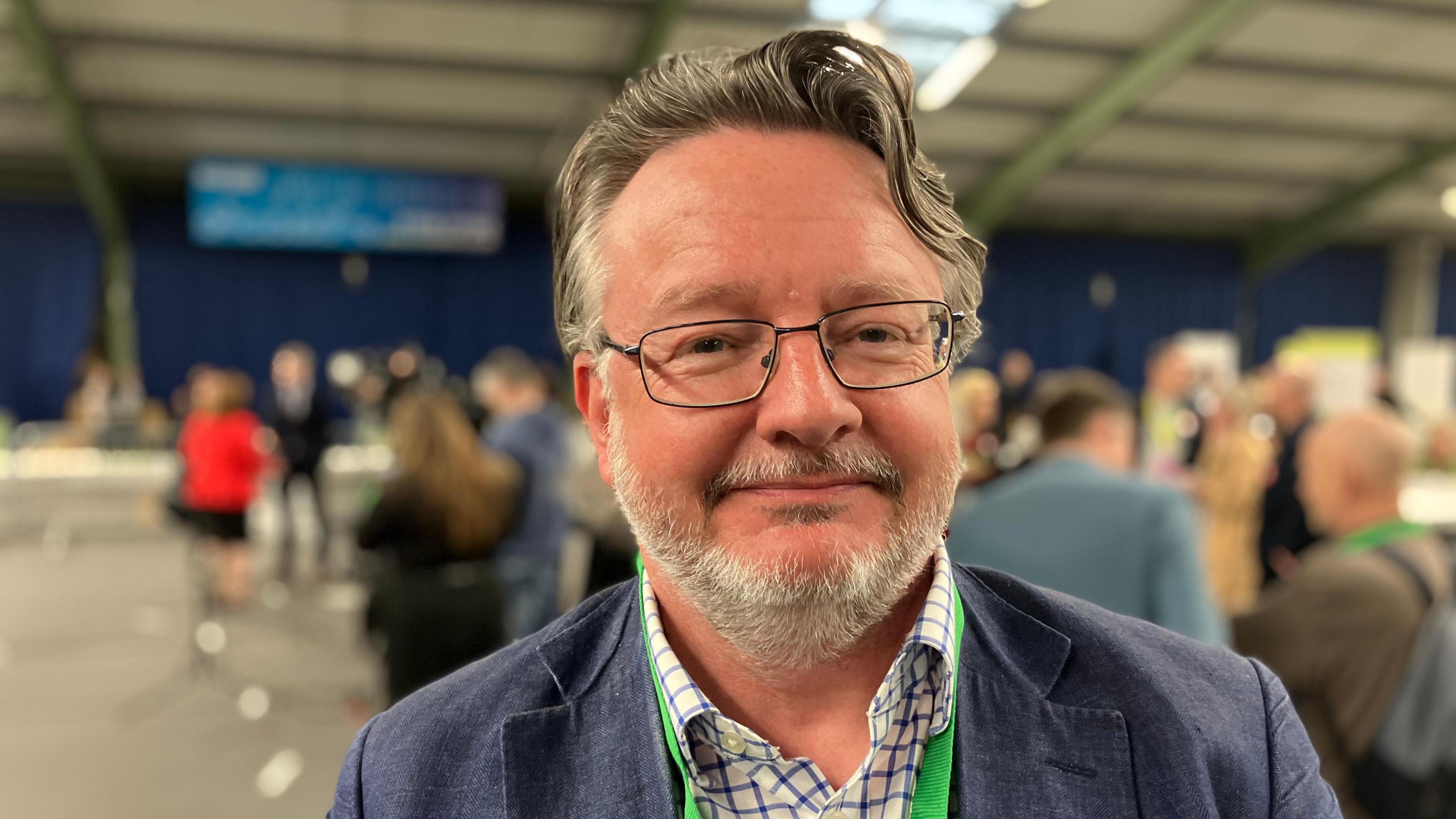 Derbyshire County Council leader Barry Lewis. He is wearing a blue jacket and a stripped shirt. He is wearing glasses and has a green lanyard around his neck.