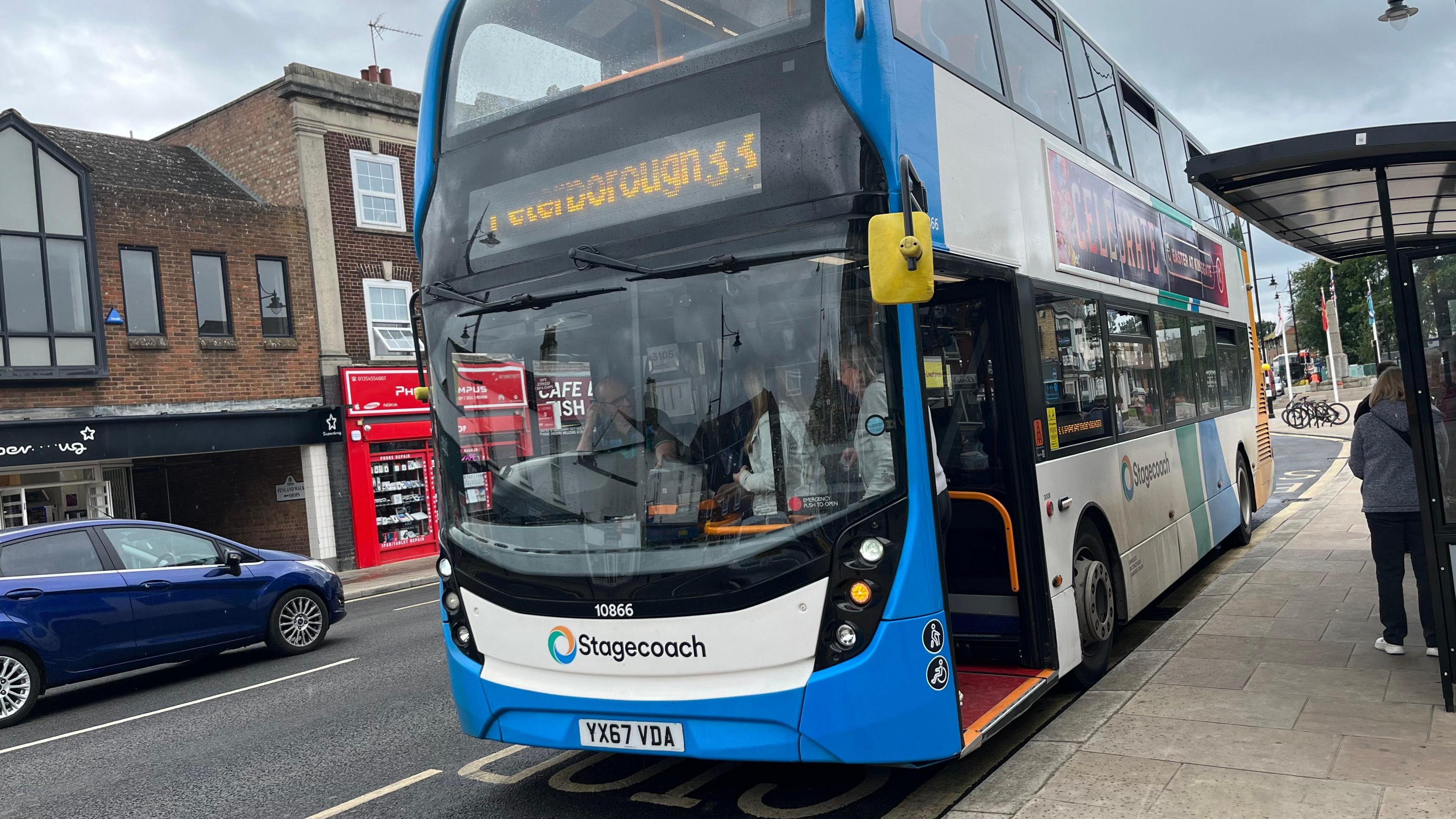 The double-decker number 33 bus standing at a bus stop in March. It has blue and white livery and Stagecoach written under its windscreen. Above the windscreen is yellow lettering saying Peterborough and 33. A couple of people can be seen standing at a bus shelter.