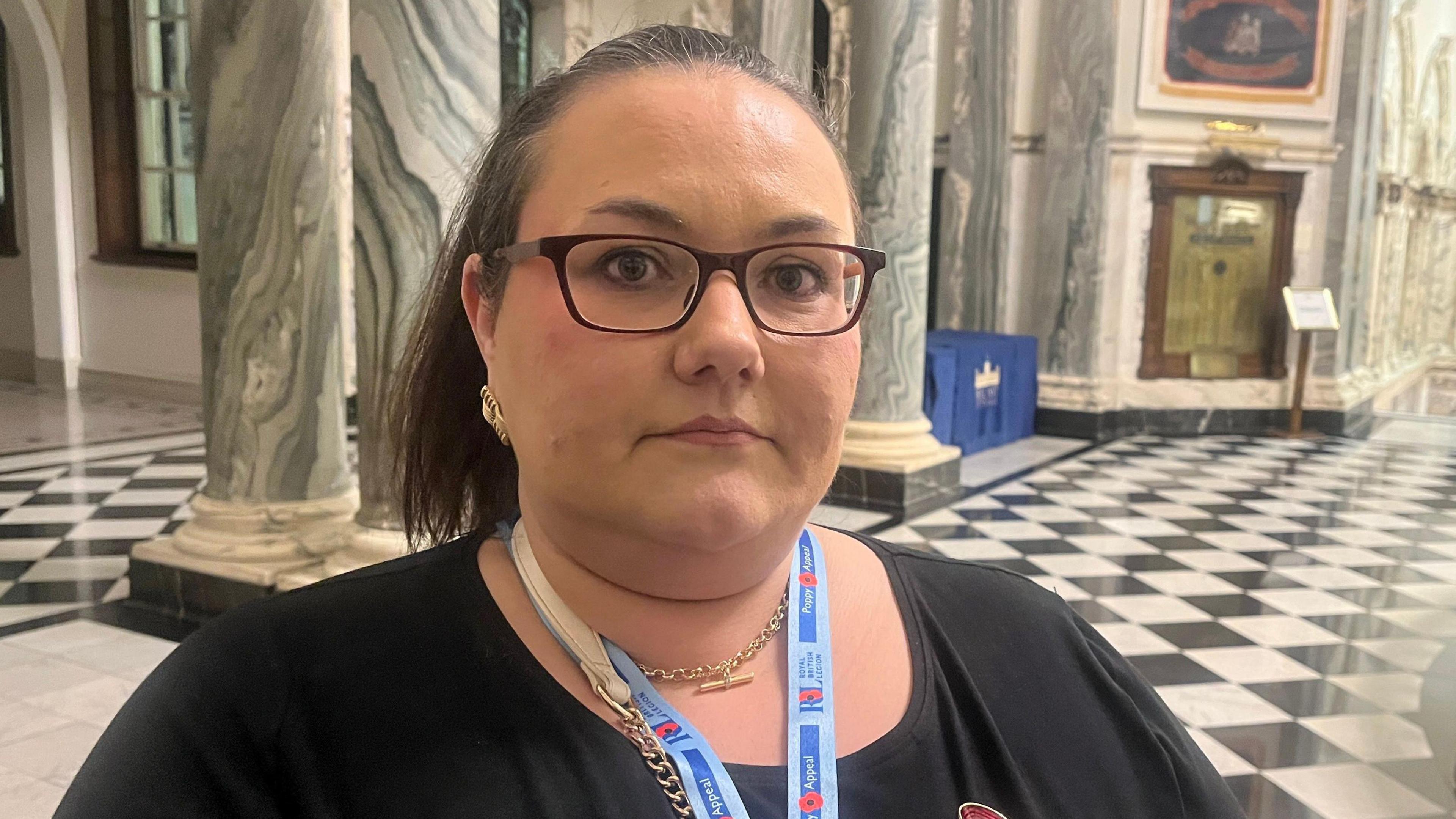 Sarah Bunting is inside city hall, which has black and white tiled floors and marbled walls with paintings, Sarahhas dark brown hair tied up, she is wearing a black top and a red poppy. She also has dark framed glasses on. 
