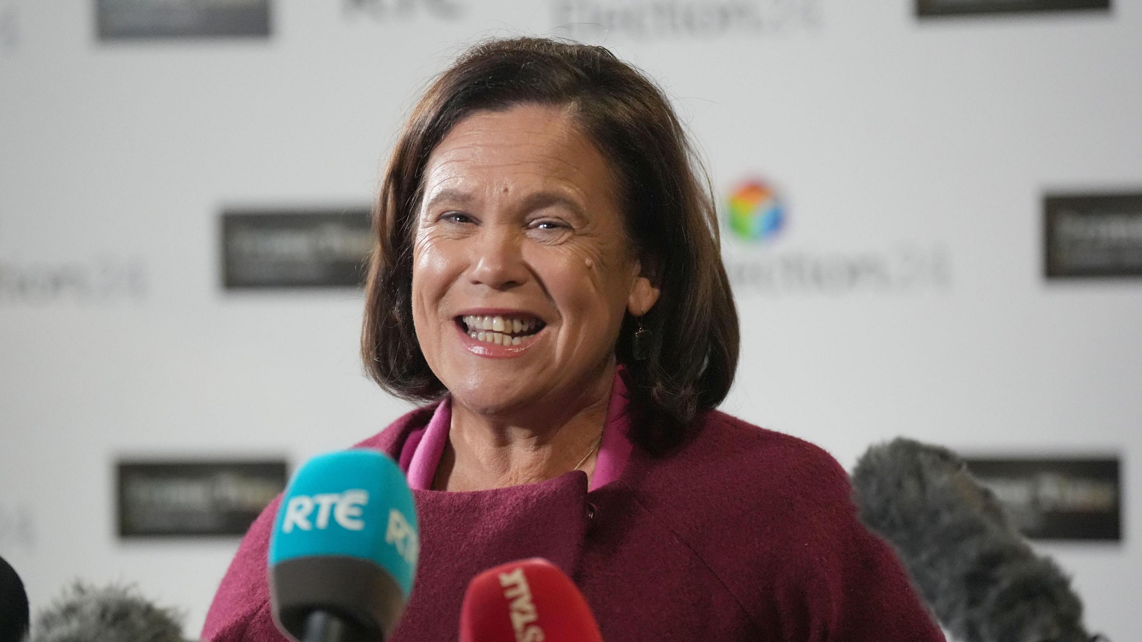 A woman with dark hair laughing in front of media microphones. She is wearing a red jumper