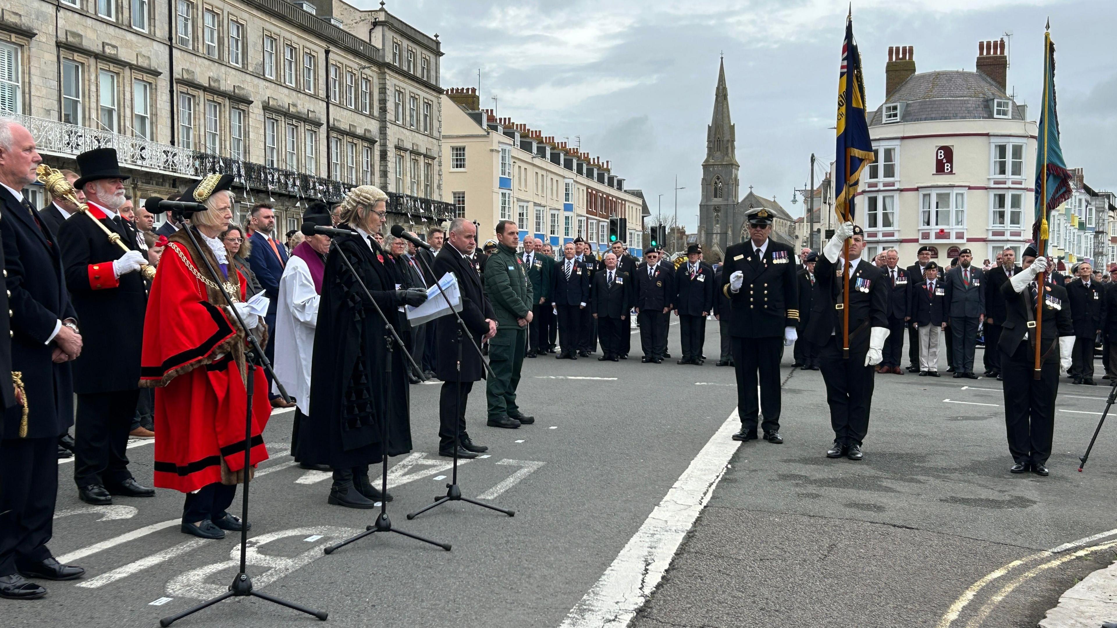 The Mayor of Weymouth, Caroline Nickinson, chaplain Father Scott Anderson, veterans and councillors during the town's Remembrance Service. It is overcast.