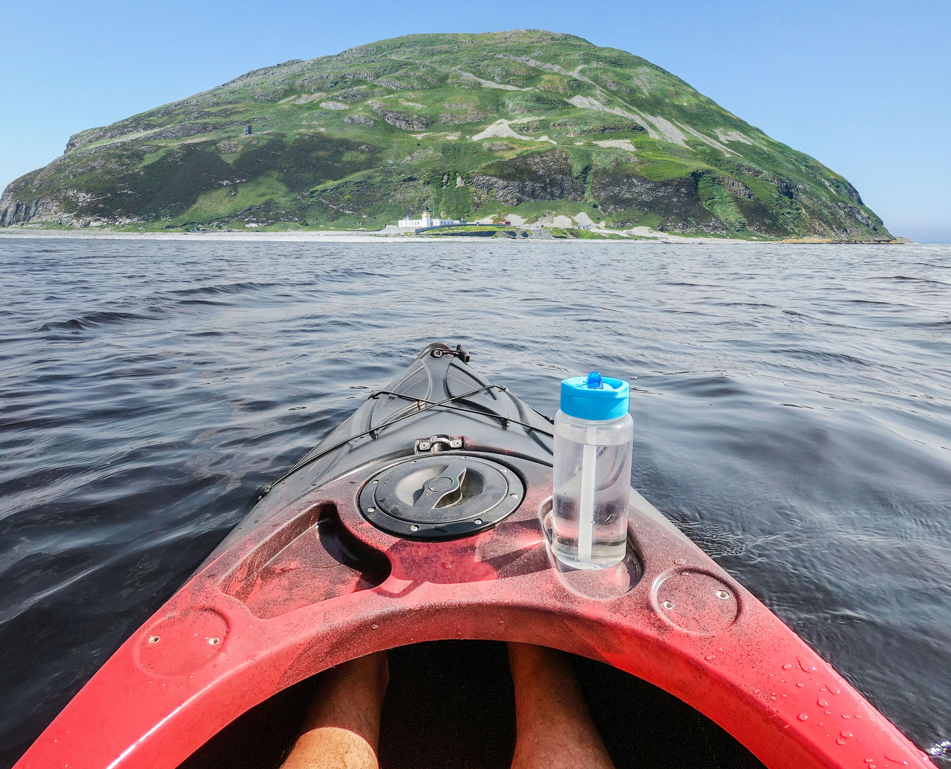Red kayak with a water bottle on the rim, floating in the water from the point of view of the kayaker with the Ailsa Craig in the distance