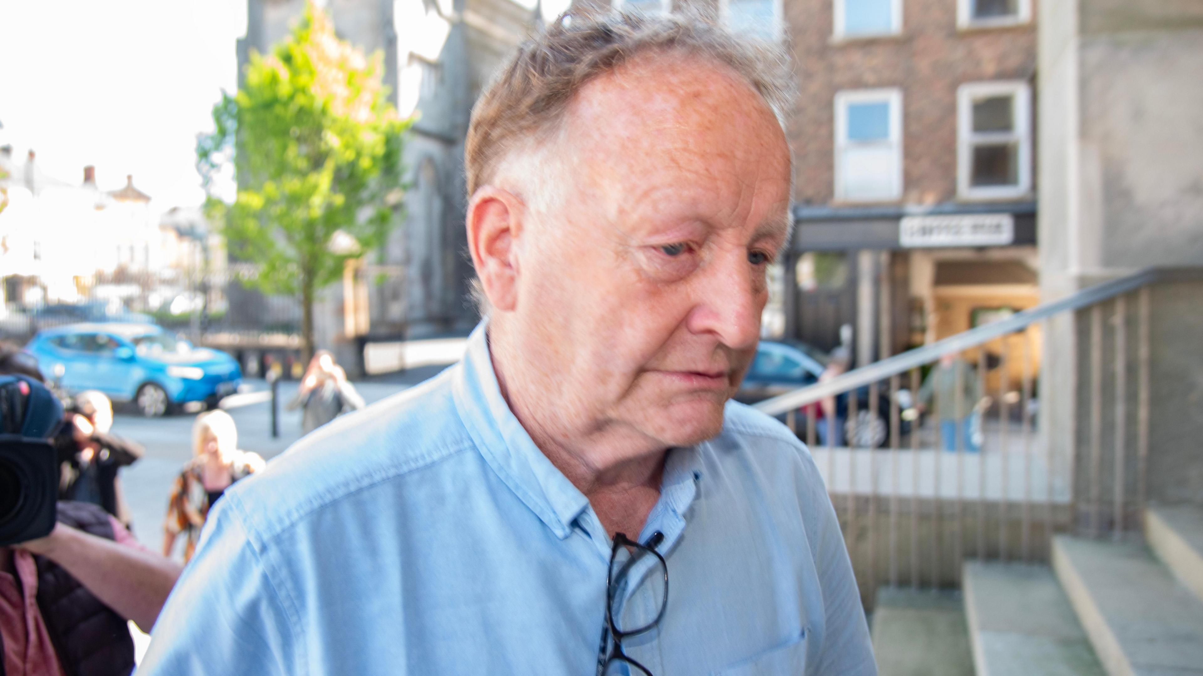 Anthony McGinn walking up concrete steps into a courtroom. The camera is close to his face as he looks away. He has messy brown and white hair. He is wearing a light blue short sleeve shirt and has a pair of black-rimmed glasses tucked into the front of his shirt.