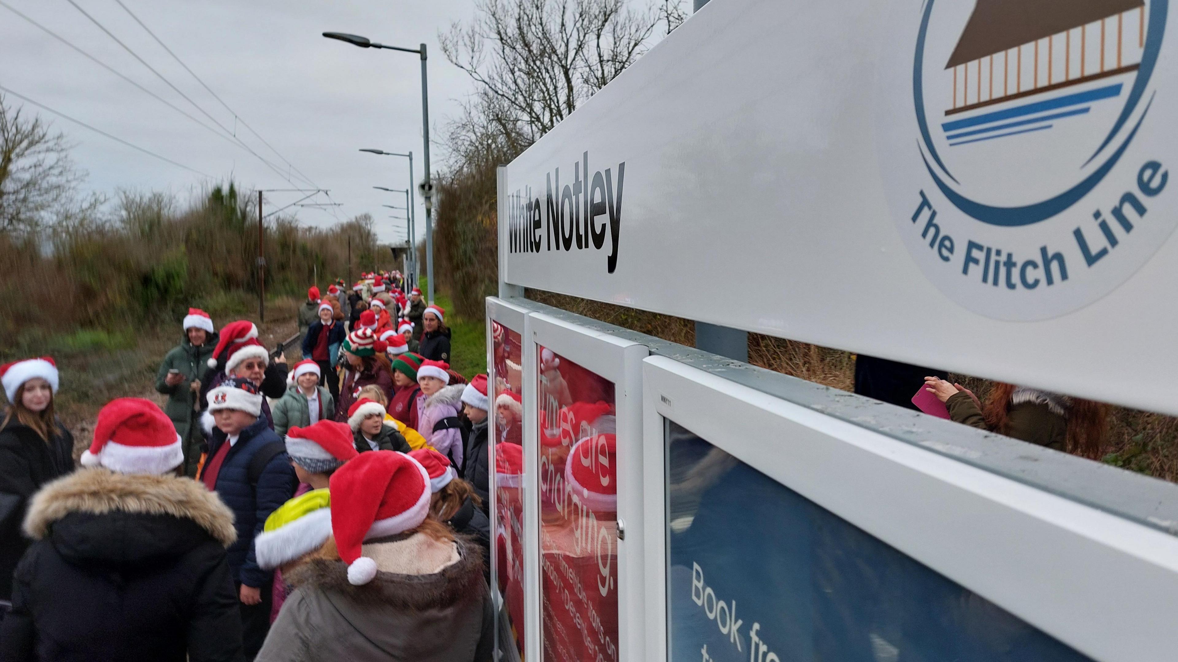 Schoolchildren wearing Santa hats stood at the White Notley train station. In the foreground is a white sign, reading White Notley, and a crest reading "The Flitch Line"