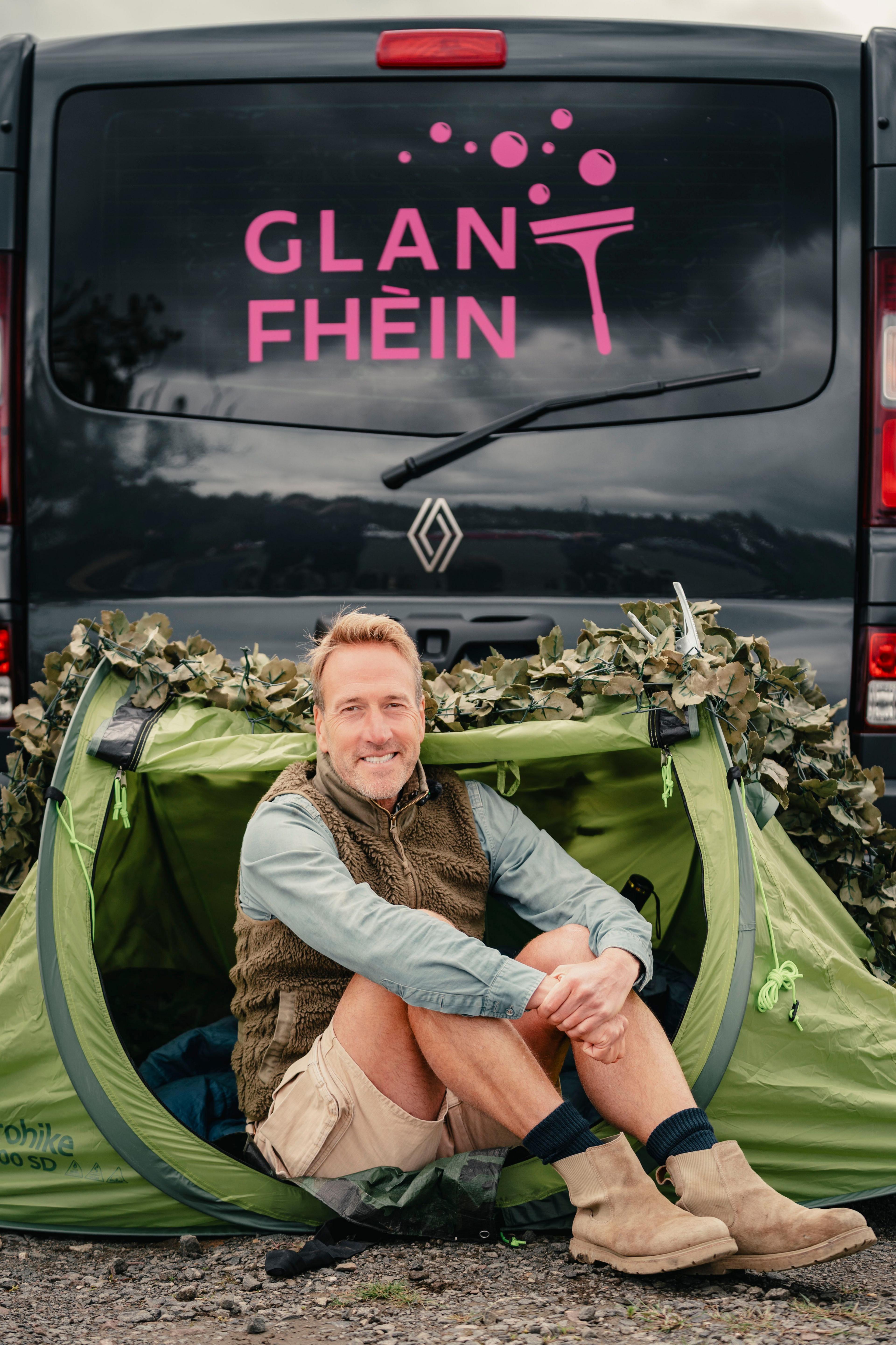 Ben Fogle na shuidhe air beulaibh teanta le bhana air a bheil Glan Fhèin sgrìobhte air a chùlaibh