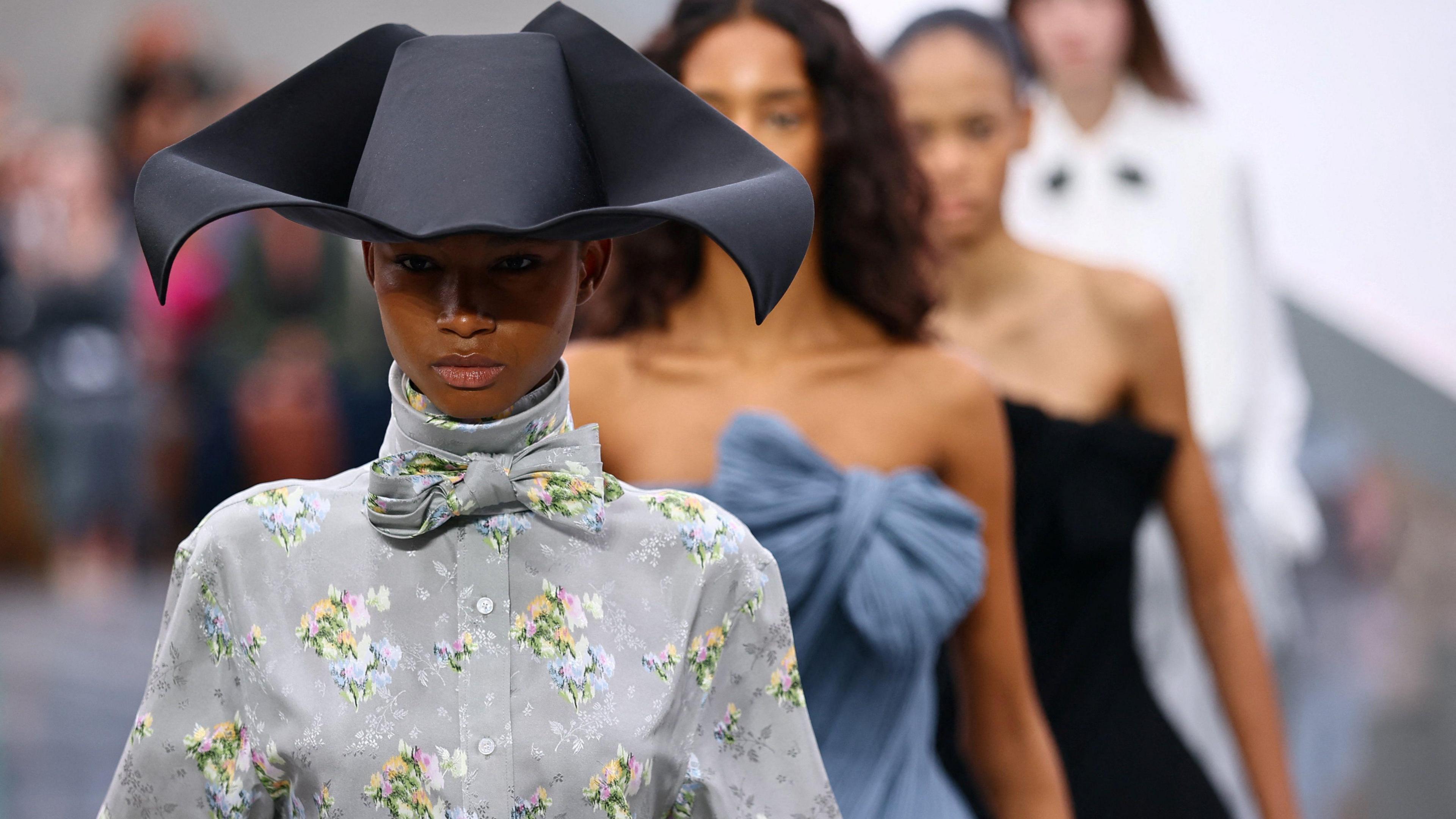 A woman in a grey shirt with floral prints. She wears a black hat. There is a line of women in dresses and shirts behind her. They are walking on a catwalk.