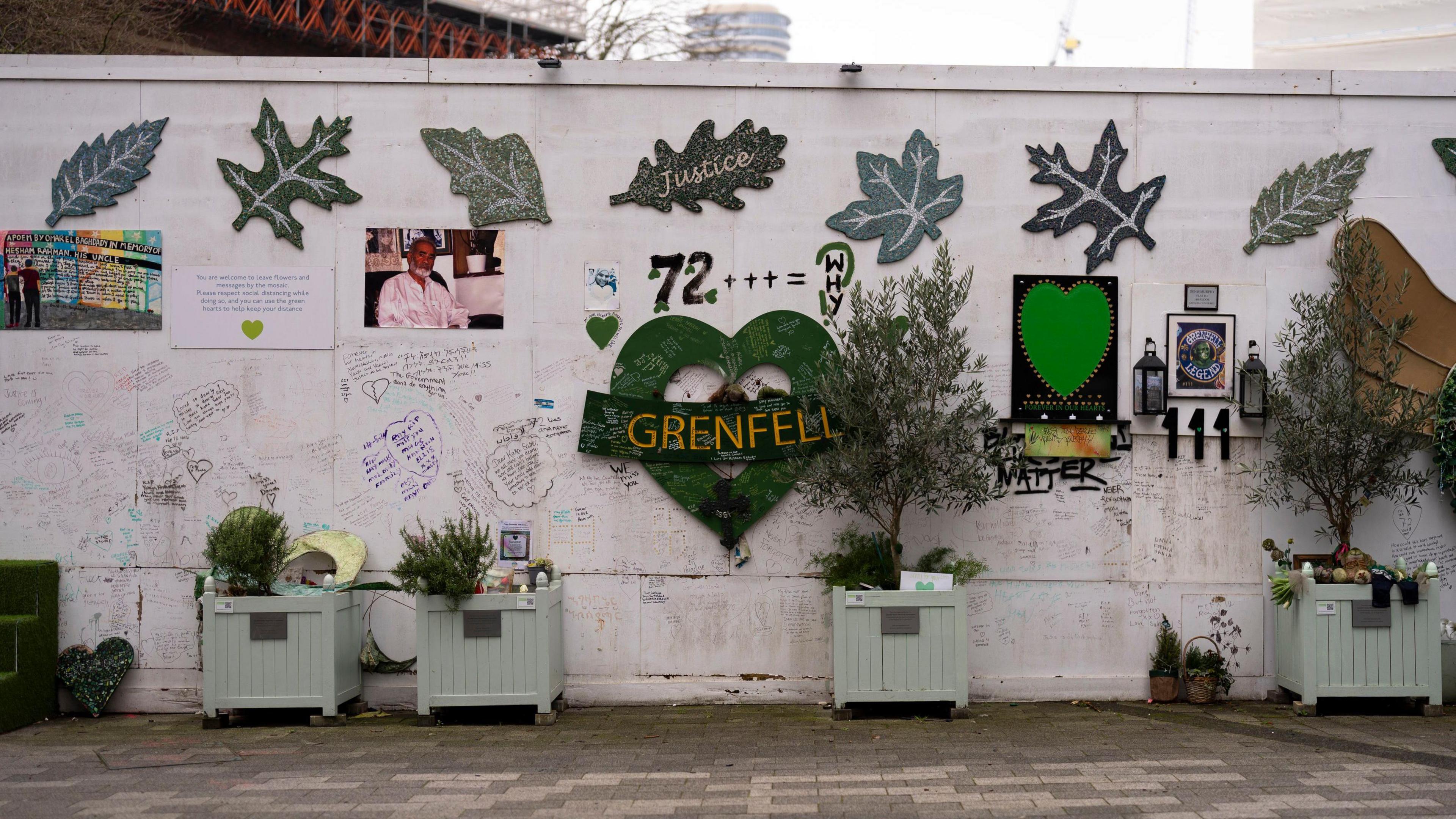 The memorial beneath Grenfell Tower, with a green heart with the words Grenfell written in the middle, the number 72 above it, alongside the words 'WHY', some pictures and large posters of leaves with the wording 'Justice'.