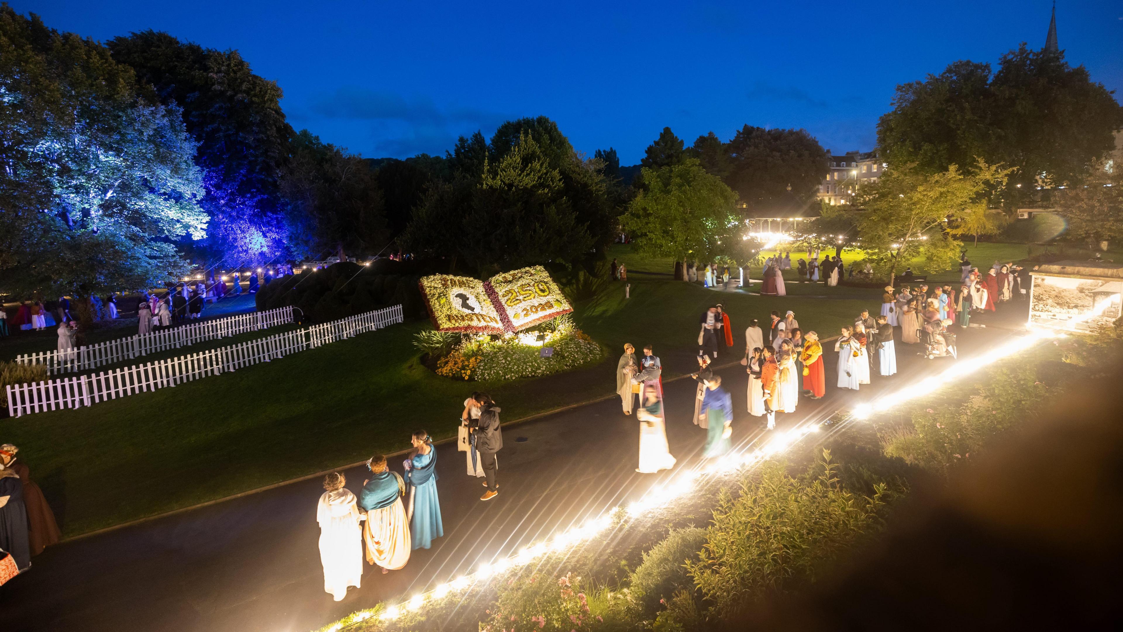 A photo of Sydney Gardens in Bath. There is a large group of people wearing regency costume as part of the Jane Austen Festival. There is a floral display depicting an open book that reads 250 2025. The silhouette of Jane Austen is also part of the display. Lights can be seen in the gardens.
