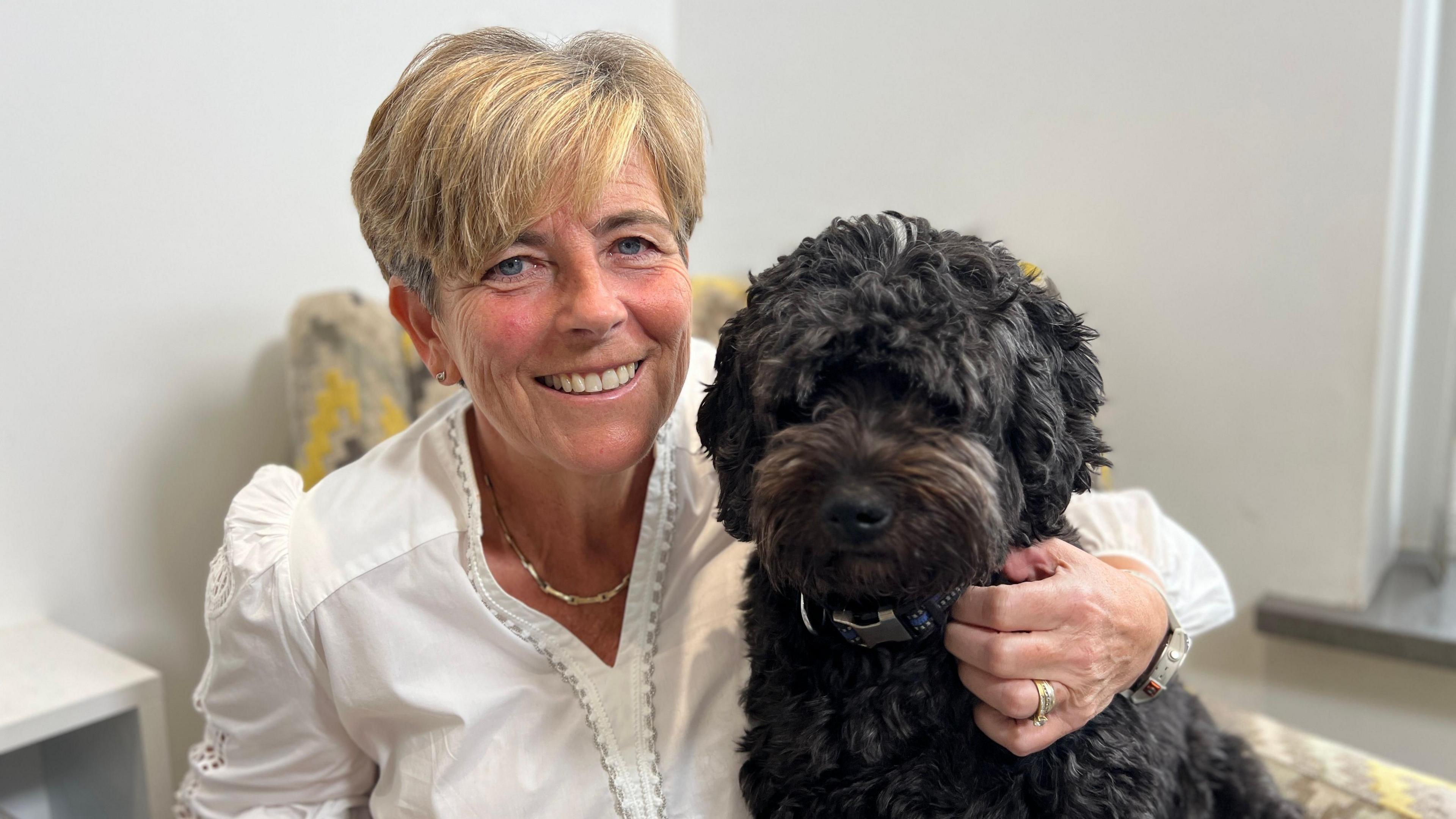 Lisa Noble smiling into the camera with her small black dog. It was taken at the same time as the first picture, with her wearing her white top.