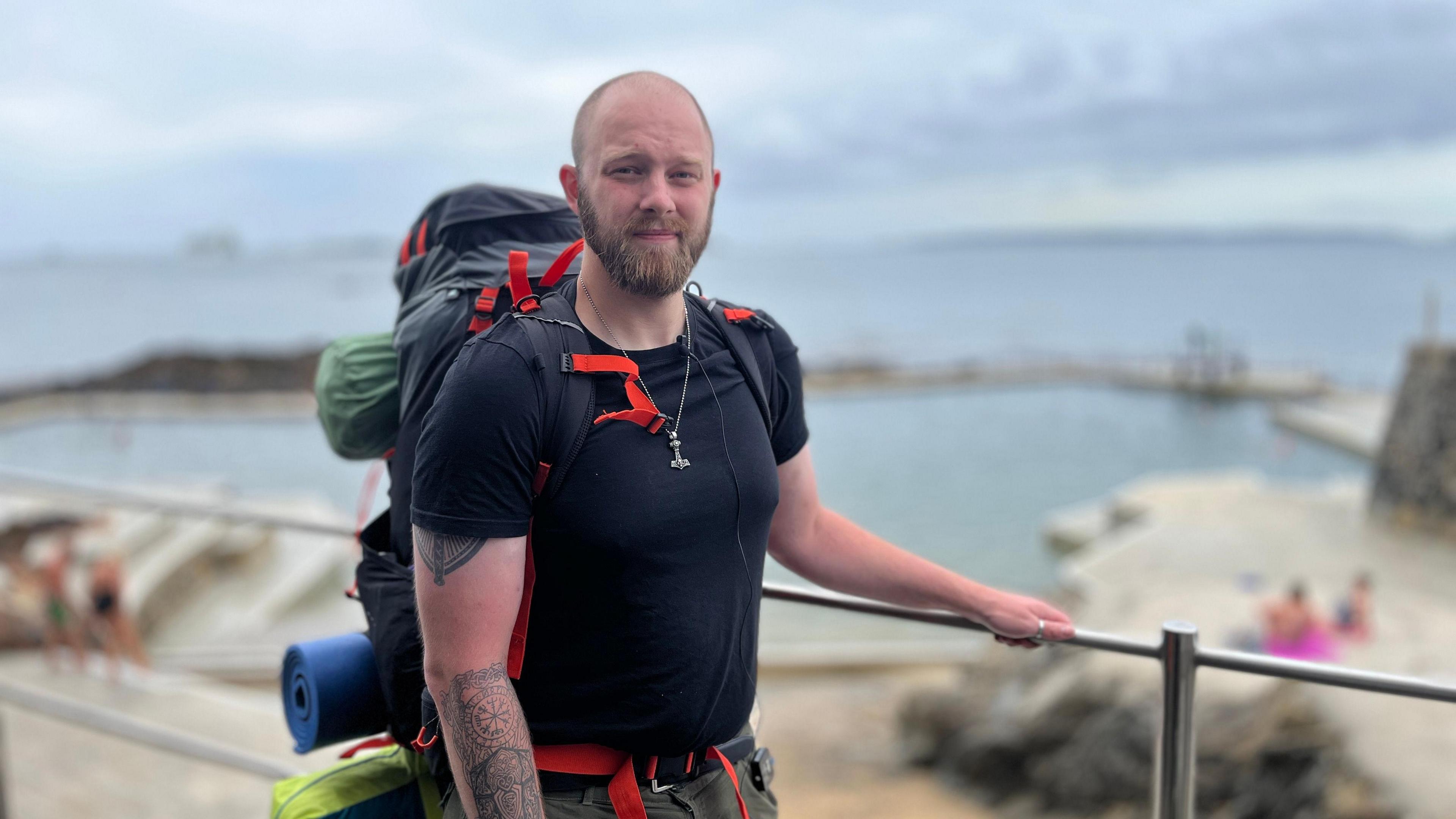 A bald, bearded man in his late 20s by some railings in front of some outdoor bathing pools. He has tattoos on his forearm, wears a black T-shirt and green shorts, and carries a large backpack.