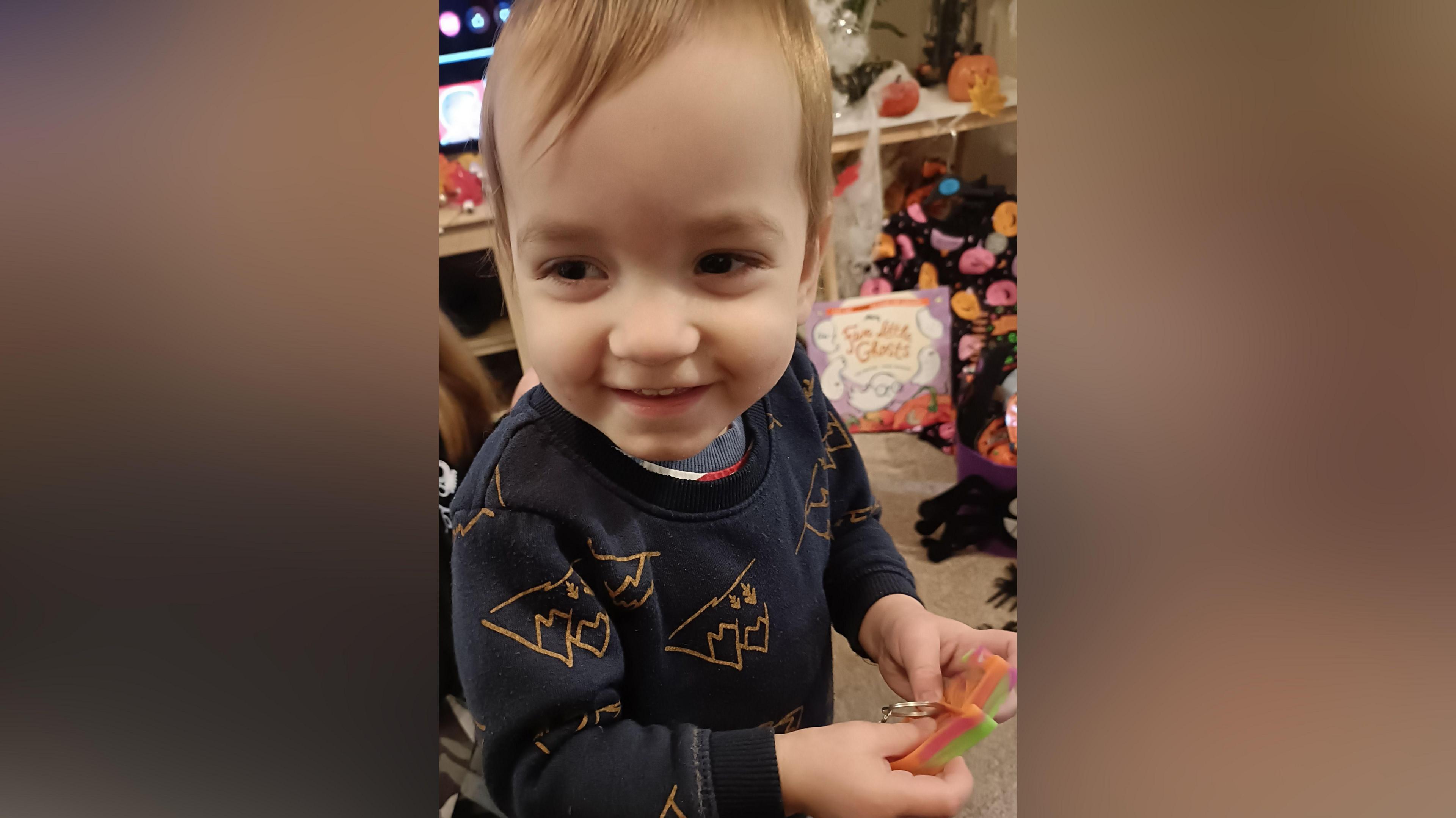 A young boy in a navy jumper with fair hair smiles as he holds a small toy in his hand