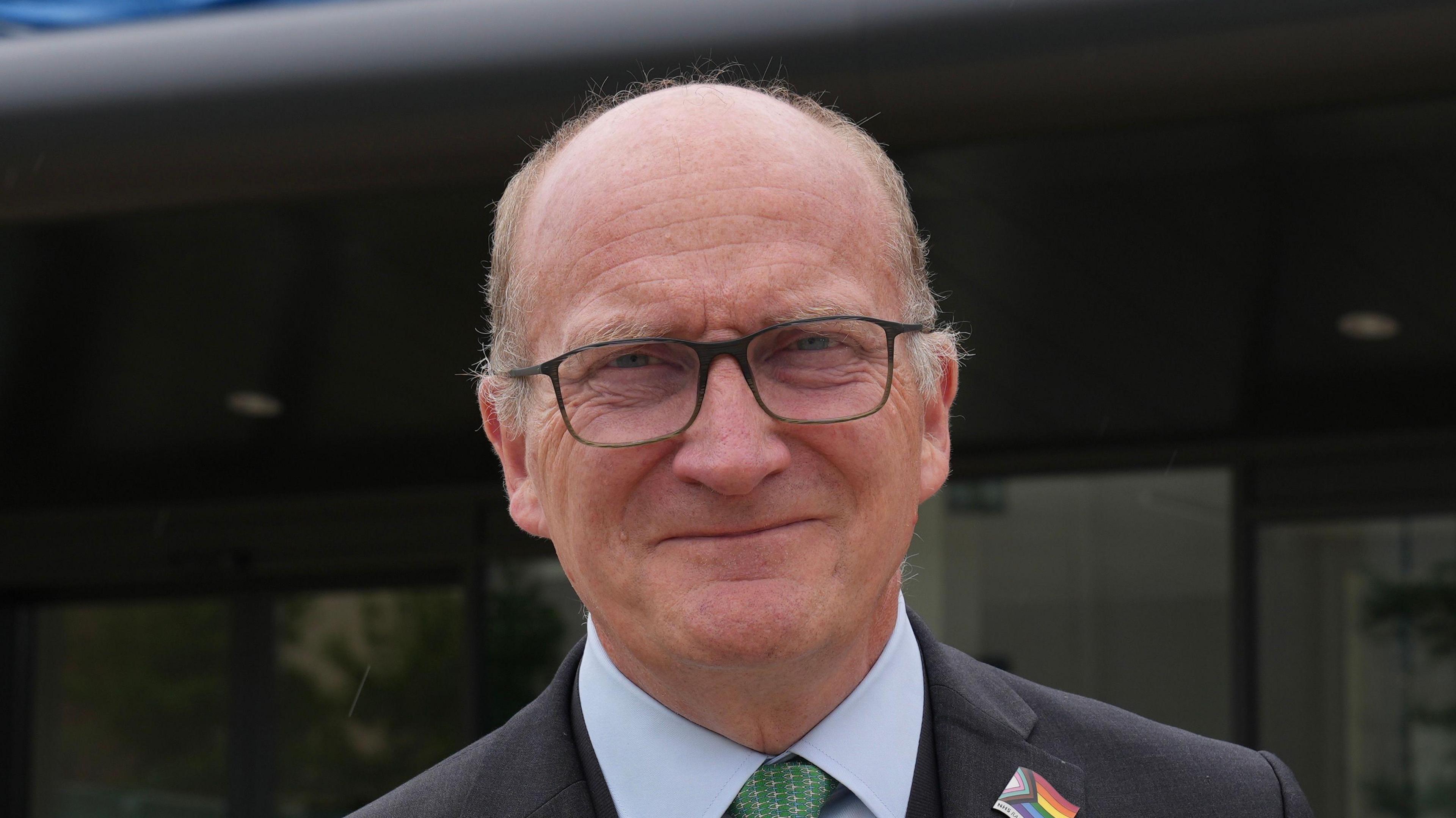 Nick Hulme smiles at the camera. He is balding with some thin grey hair around the edge of his head. He wears glasses and a black suit with a white suit and green tie. A Pride flag badge is pinned to his collar.